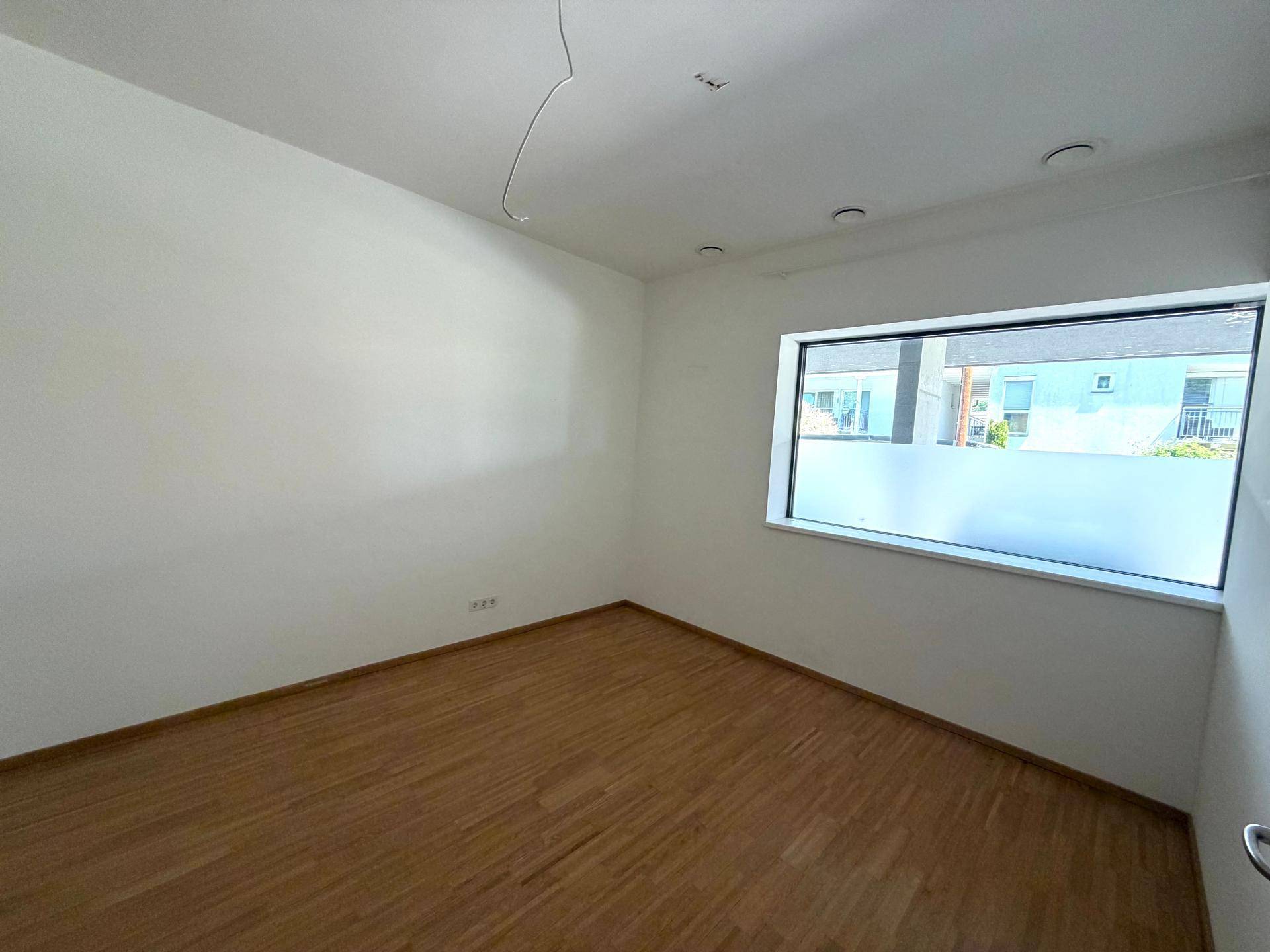 Leerer Raum mit hellem Holzfußboden, weißen Wänden und einem breiten Fenster mit Sichtschutz.
