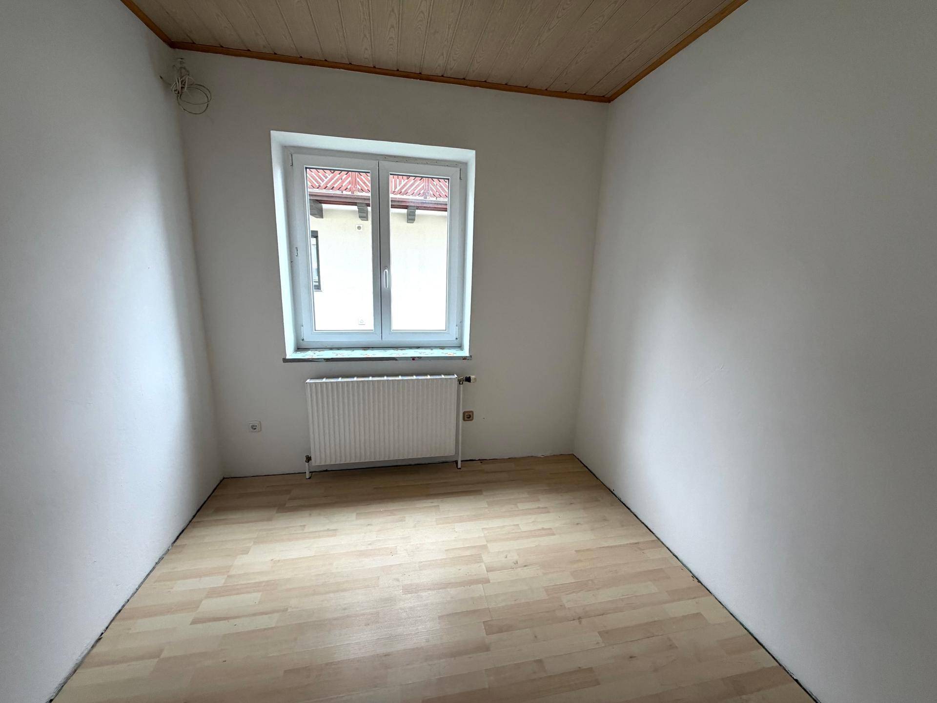 Kleines, helles Zimmer mit Holzboden, weißen Wänden und Heizkörper unter dem Fenster.