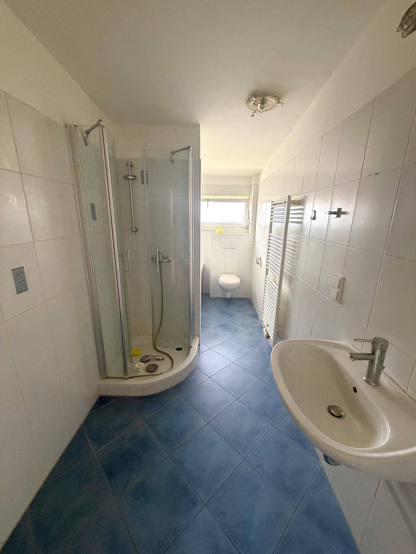 Badezimmer mit Dusche, Waschbecken, WC und blauen Bodenfliesen in gutem Zustand.