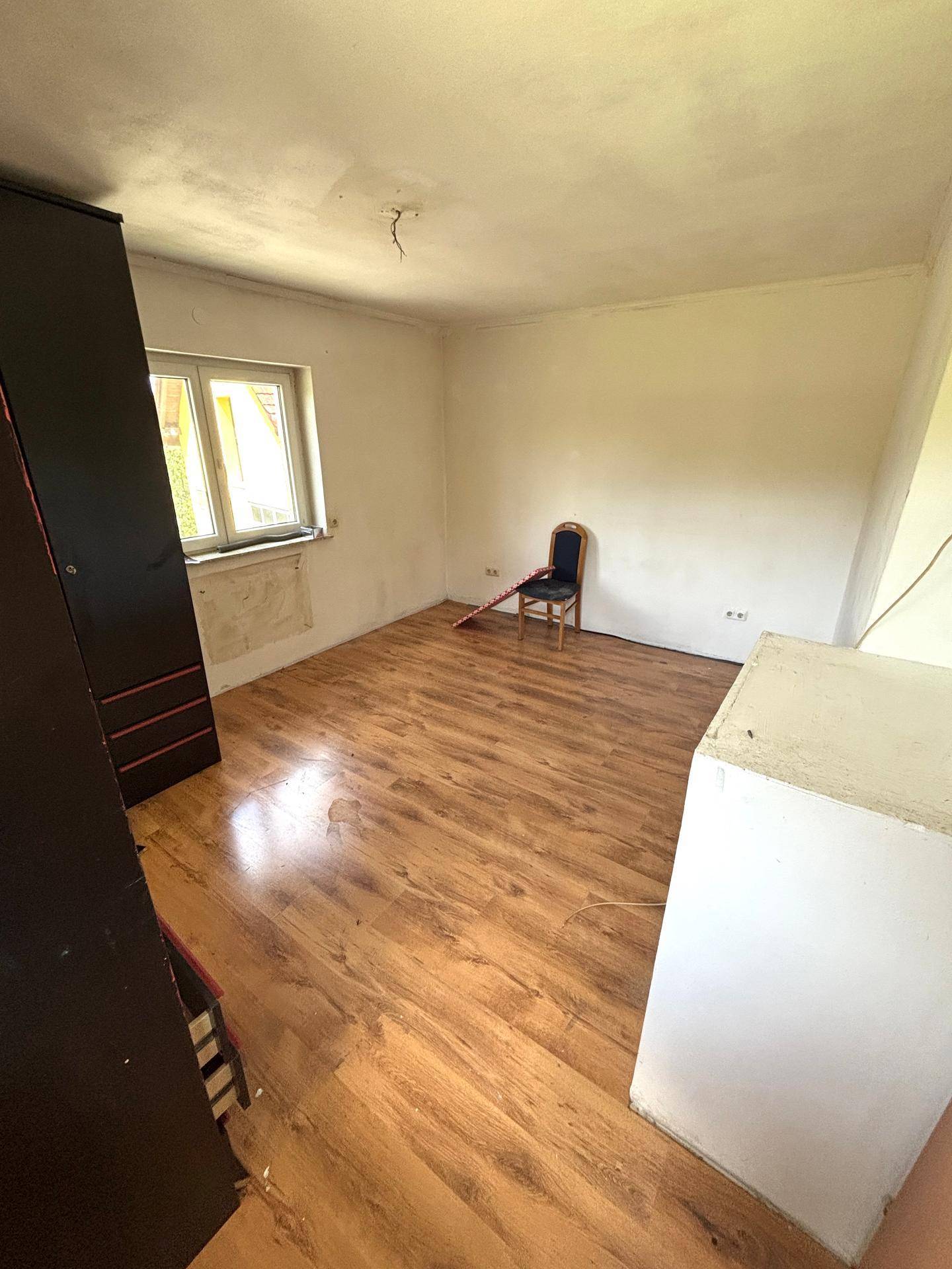 Helles Zimmer mit Laminatboden, weißen Wänden und einem Fenster, ein dunkler Schrank ist sichtbar.