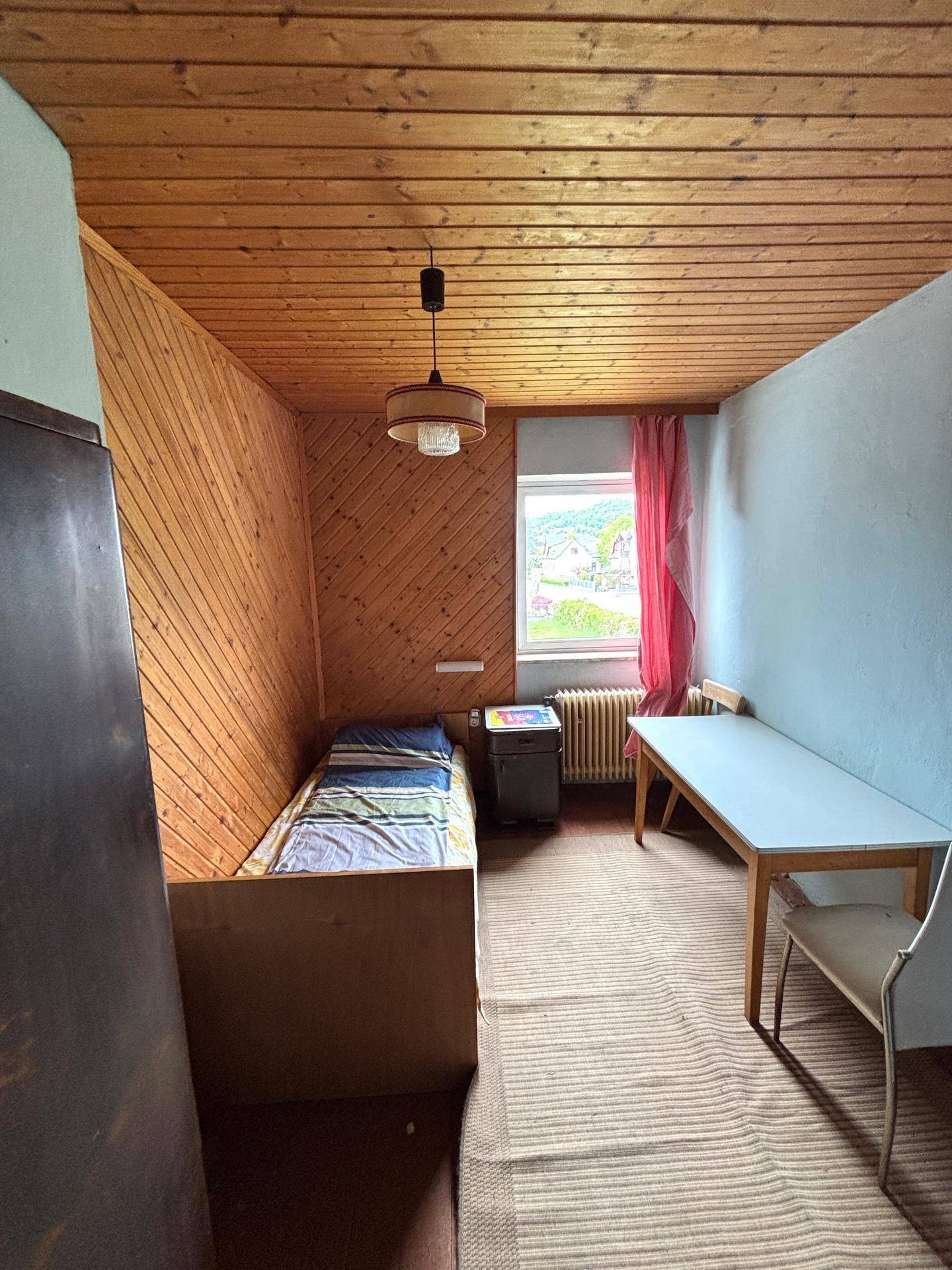Schlafzimmer mit Holzwänden, Holzdecke und Einzelbett, hell durch Fenster.