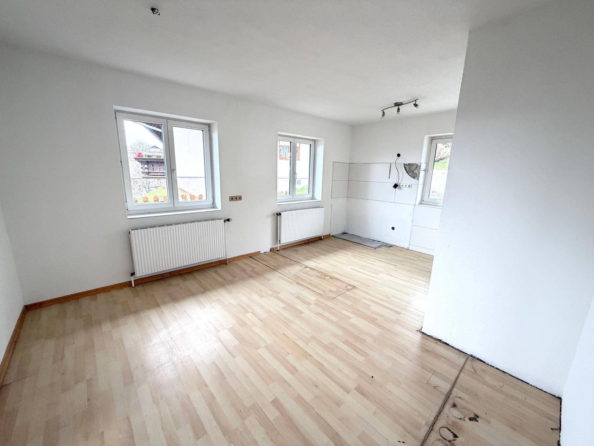 Leerer Raum mit hellem Holzboden und Fenstern, vorbereitet für eine Kücheninstallation, mit Heizkörpern.