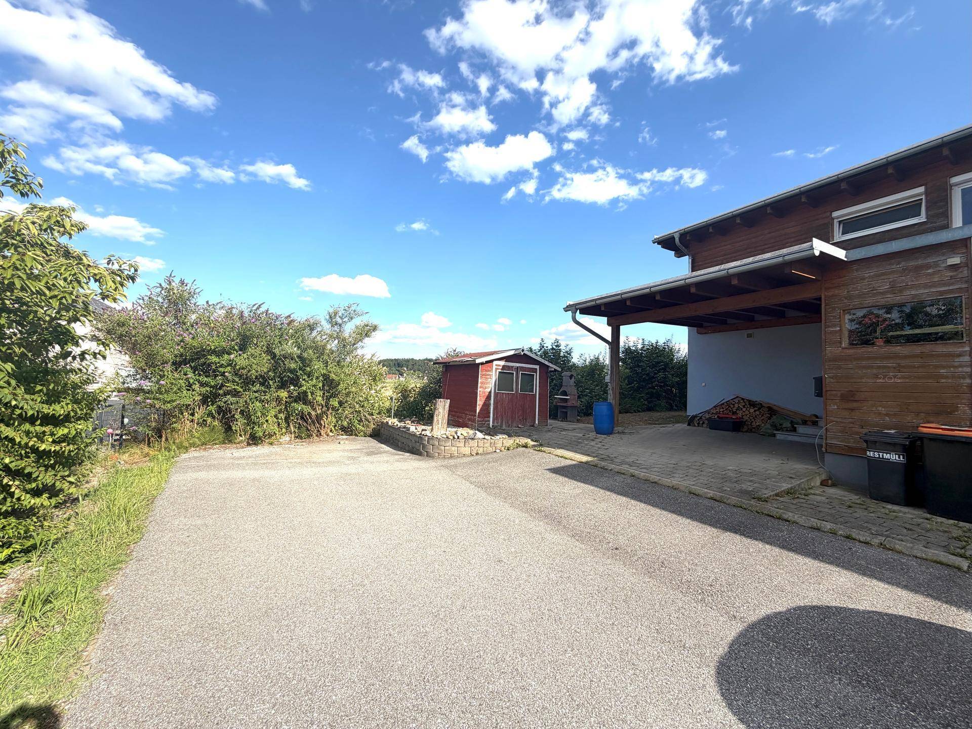 Zufahrt zum Haus mit Carport und Gartenhaus im Hintergrund unter blauem Himmel.