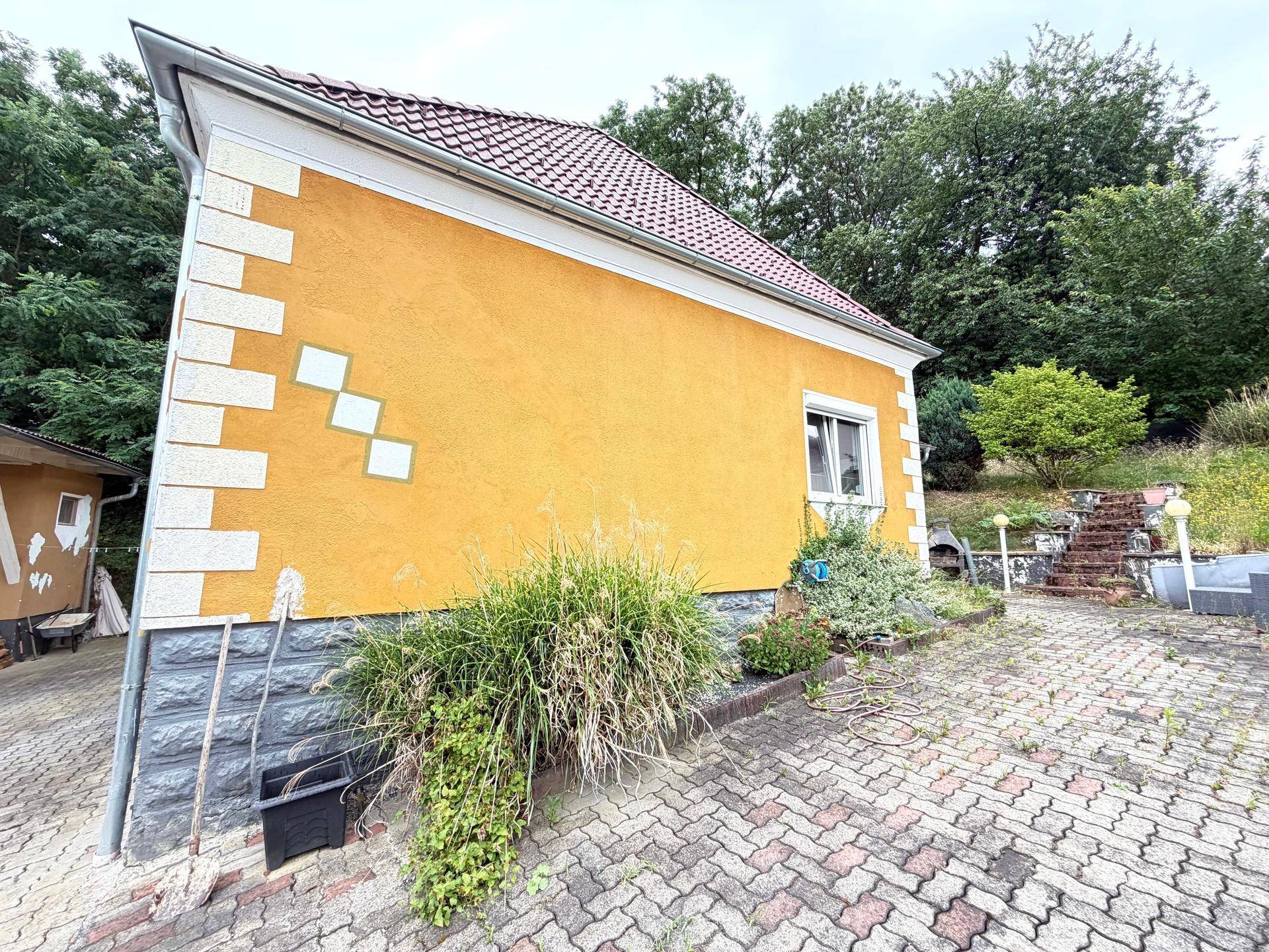 Seitenansicht eines Hauses mit gelber Fassade, roten Dachziegeln und gepflastertem Vorplatz.