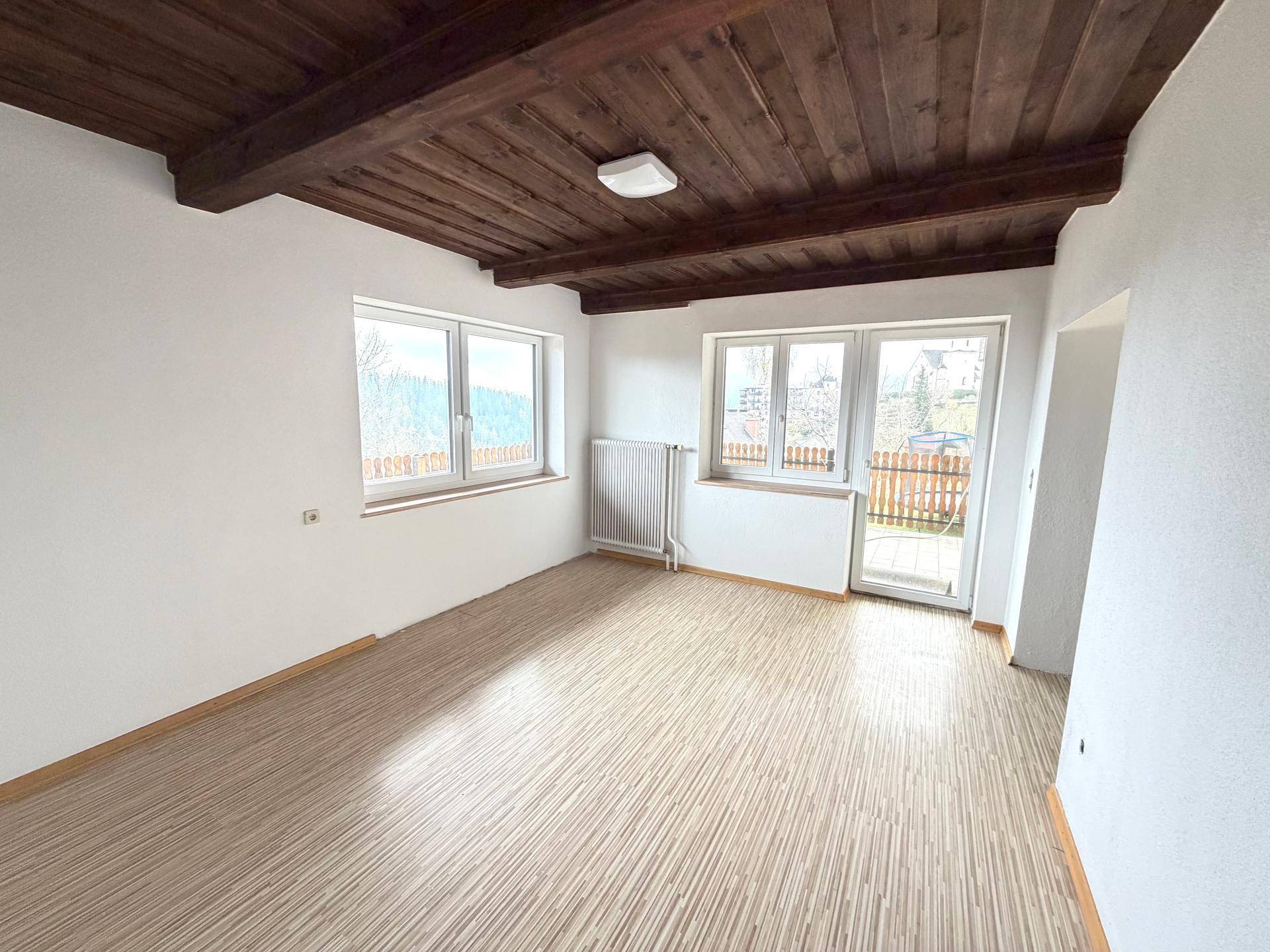 Gemütliches Zimmer mit Holzbalkendecke, hellem Boden und weitem Blick ins Grüne durch große Fenster.
