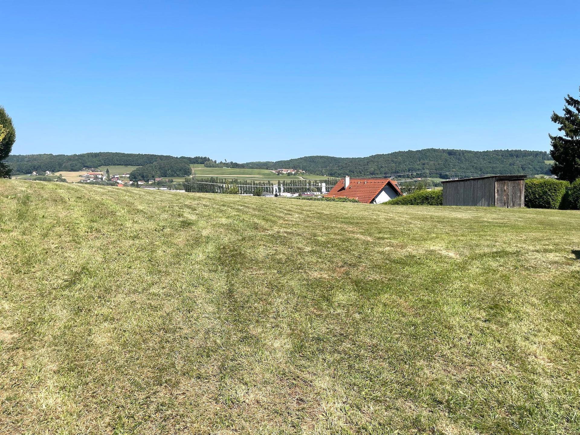 Weitläufige grüne Fläche mit Blick auf die sanfte Hügellandschaft und einige Dächer in der Ferne.