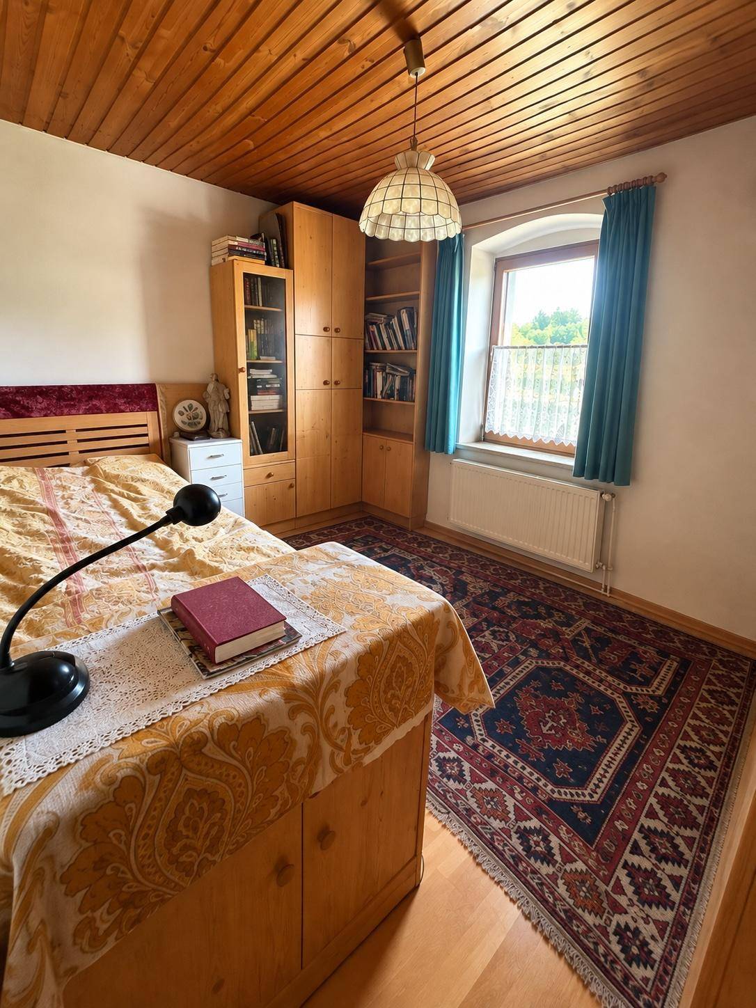 Schlafzimmer mit Holzbett, Teppich und Holzdecke, durch das Fenster fällt Tageslicht herein.