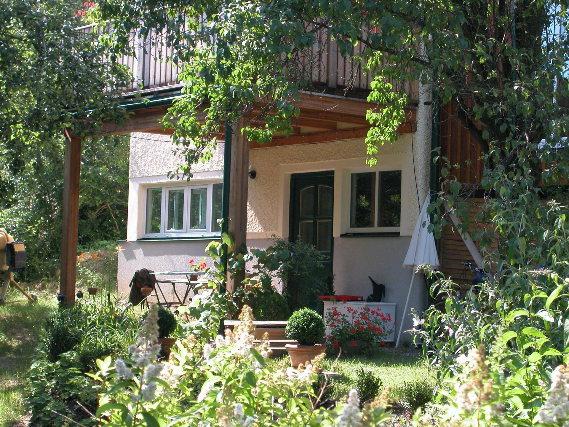 Überdachte Terrasse am Haus, umgeben von blühenden Pflanzen, ideal für entspannte Stunden im Freien.