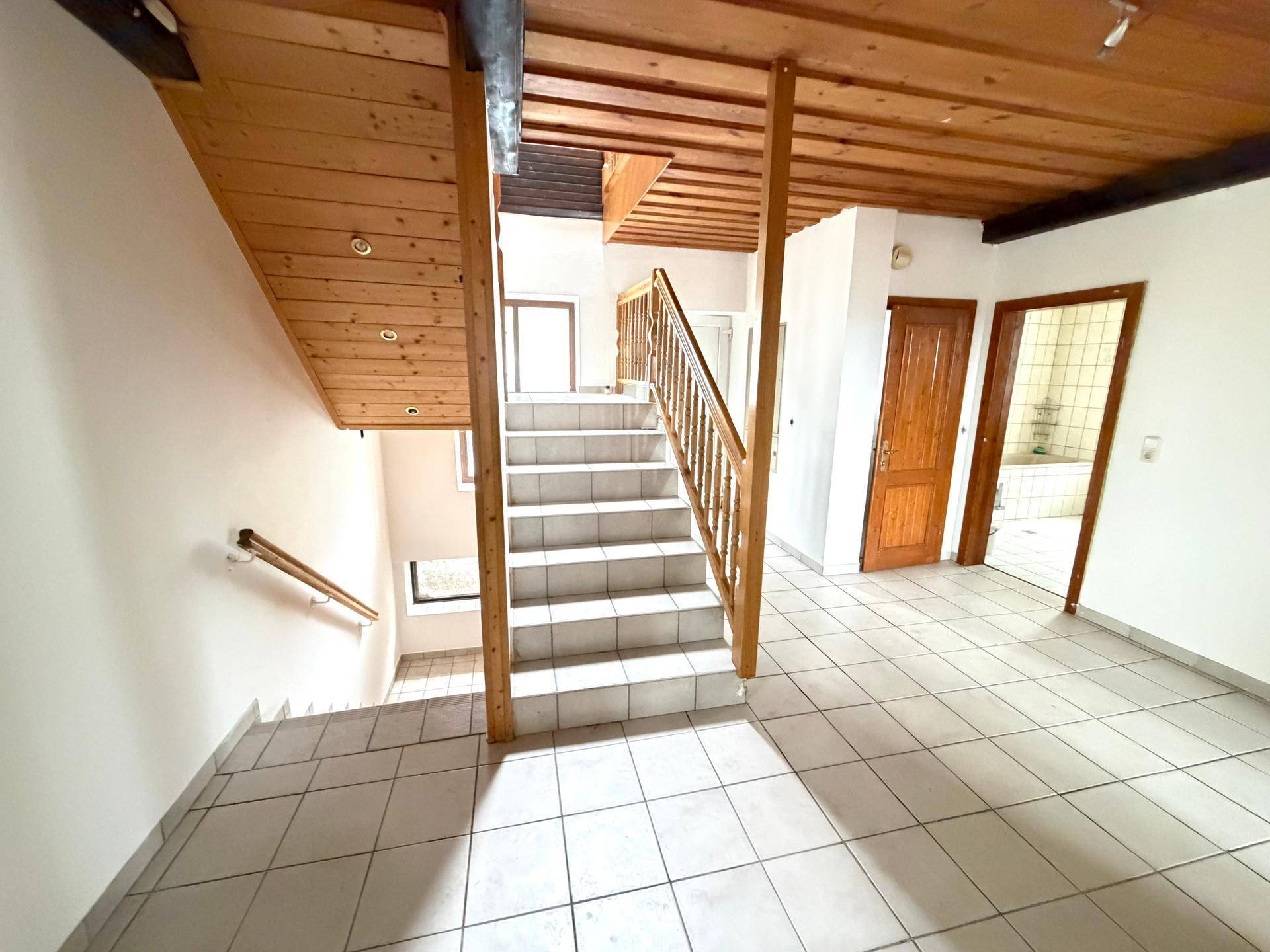 Treppenhaus mit Fliesenboden, Holztreppe und sichtbaren Holzbalken an der Decke, verbindet die Etagen.
