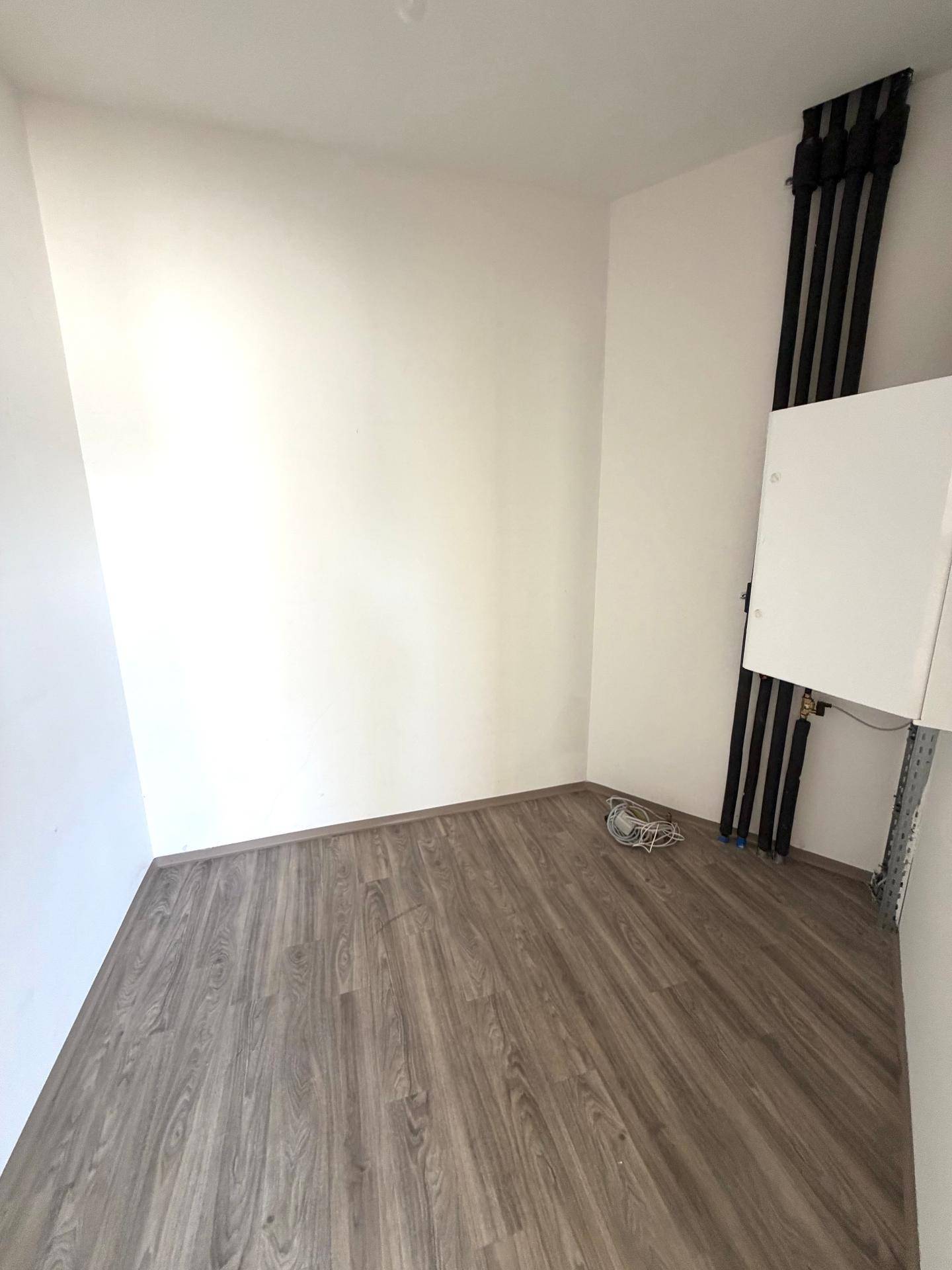 Kleiner Raum mit grauem Laminatboden, weißen Wänden und sichtbaren Installationen.
