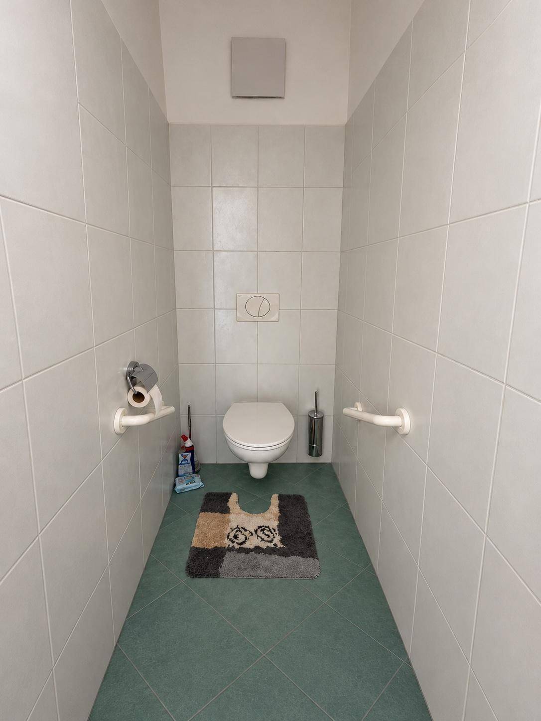 Separates WC mit hellen Fliesen, wandhängender Toilette und Haltegriffen für Komfort.