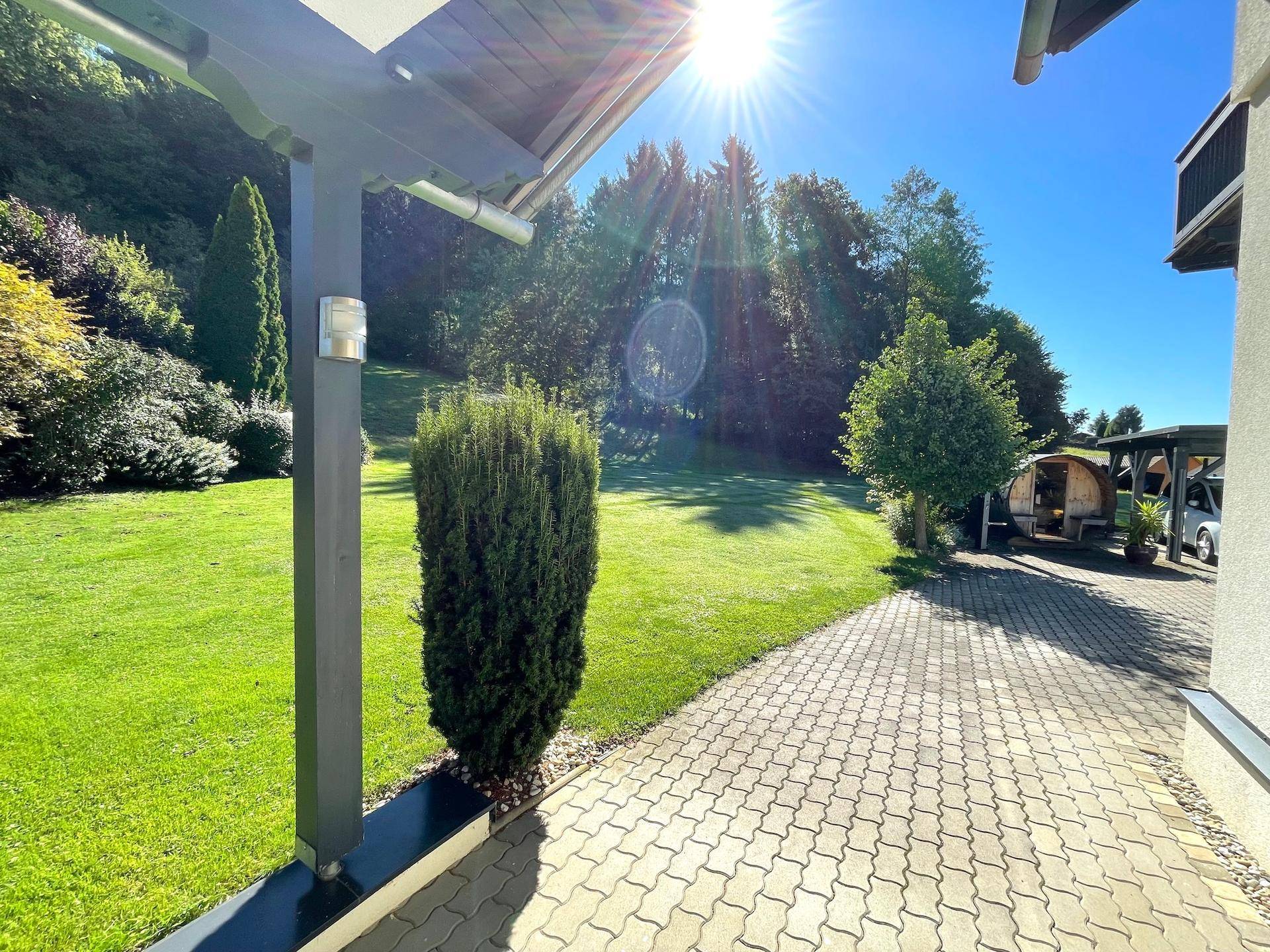 Gepflasterter Bereich neben dem Haus mit Blick auf den Garten und eine Fasssauna im Hintergrund.