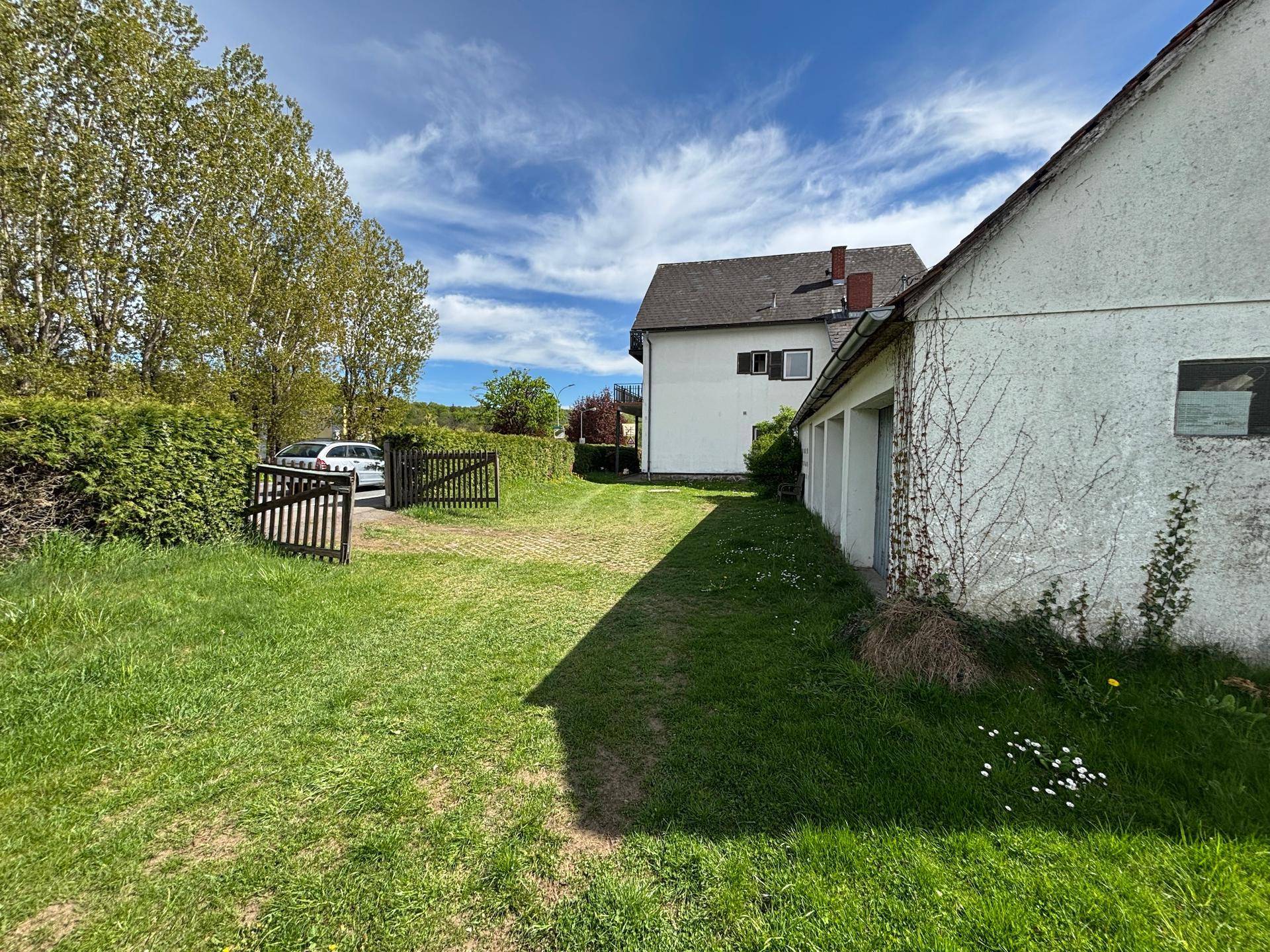 Großer Gartenbereich mit Rasen, Zufahrt und Holztor, umgeben von Hecken und Bäumen.