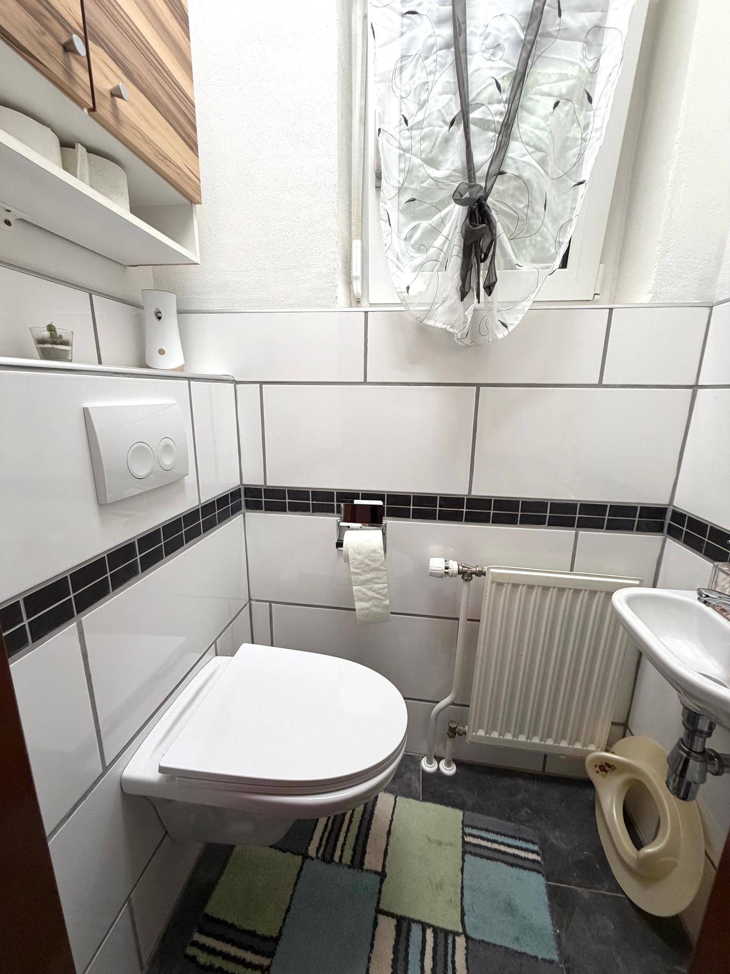 Kleines Gäste-WC mit Wand-WC, Waschbecken und schwarz-weißen Fliesen, hell durch ein Fenster.