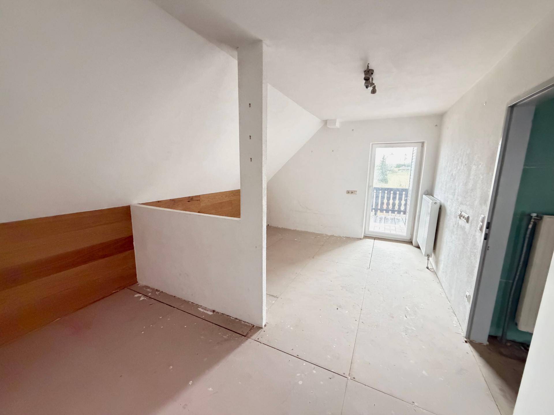 Dachgeschosszimmer im Ausbau mit schräger Decke, Holzelementen und Zugang zu einem Balkon.