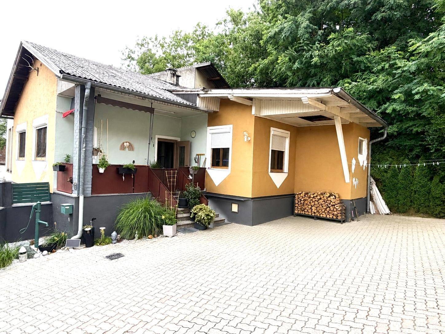 Gelbes Haus mit gepflasterter Einfahrt und Holzstapel, umgeben von Bäumen.