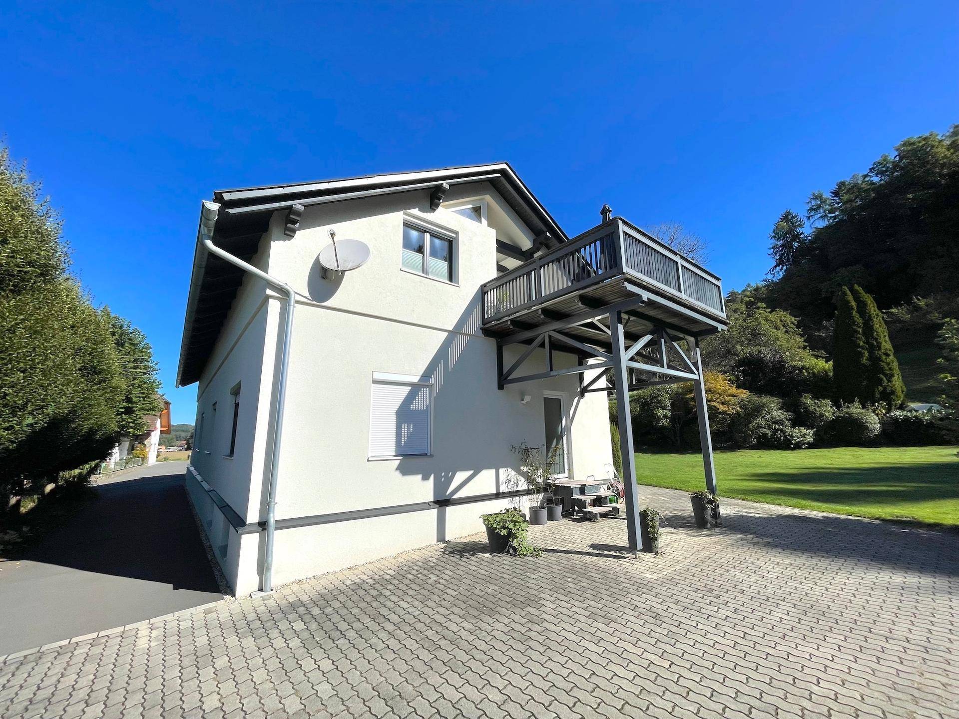 Seitenansicht des Hauses mit einem erhöhten Balkon auf Stützen und einem gepflasterten Vorplatz.