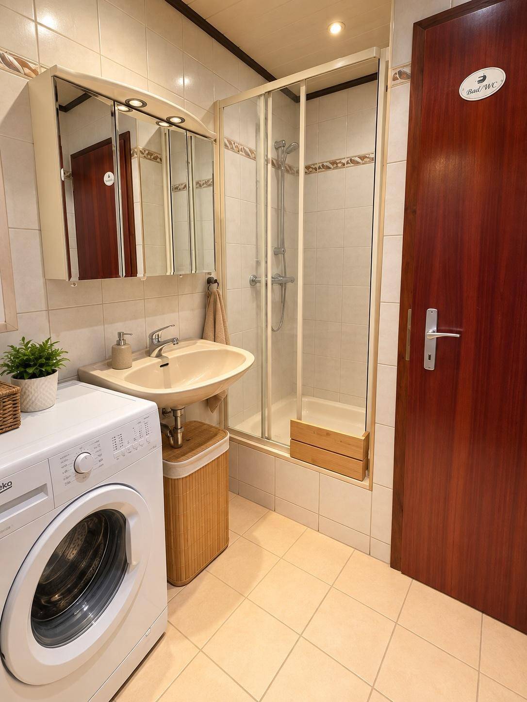 Badezimmer mit Dusche, Waschbecken, Waschmaschine und Spiegelschrank, hell gefliest.