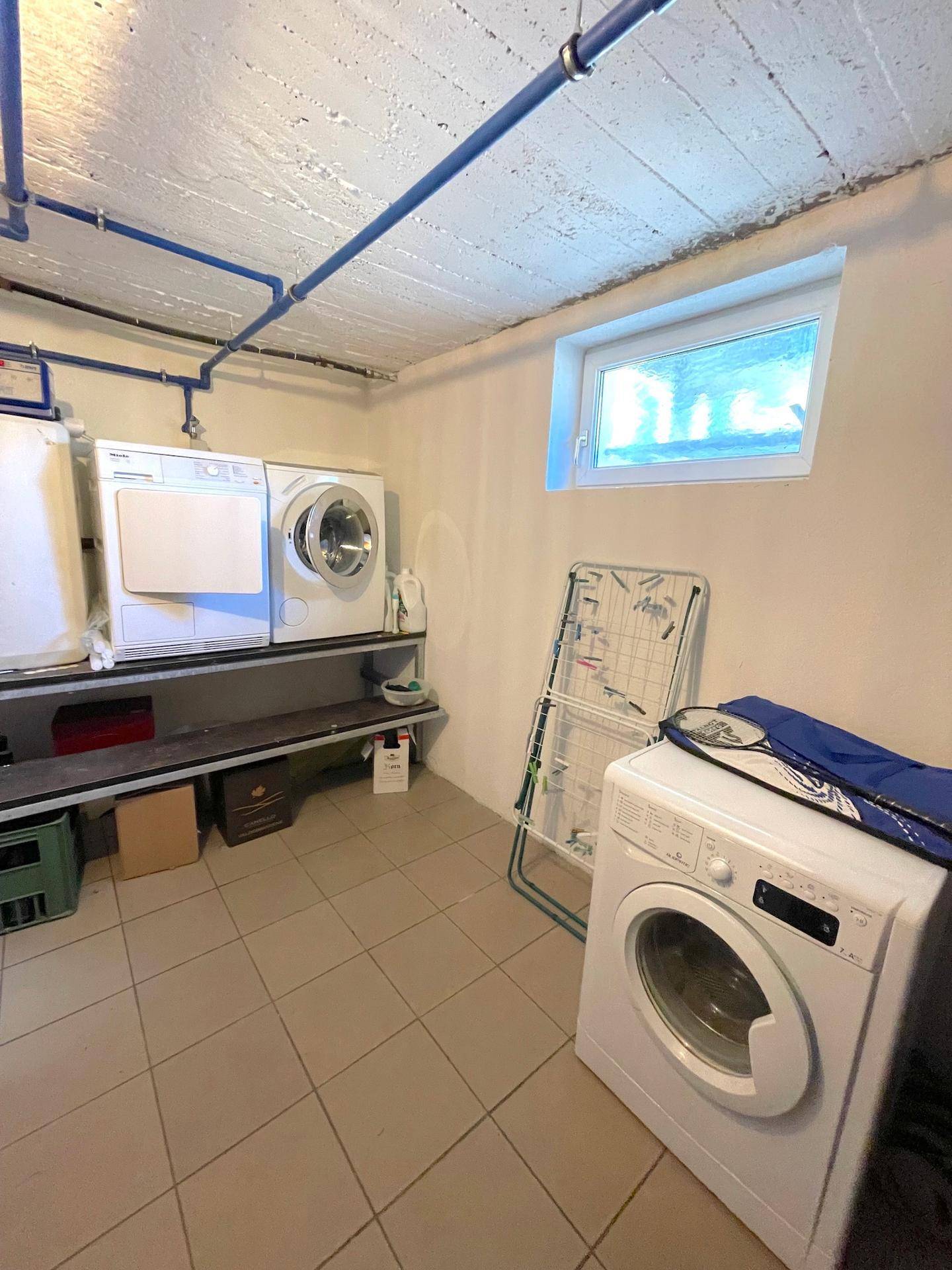 Waschküche im Keller mit Waschmaschinen, Trockner und einem kleinen Fenster für Belüftung.