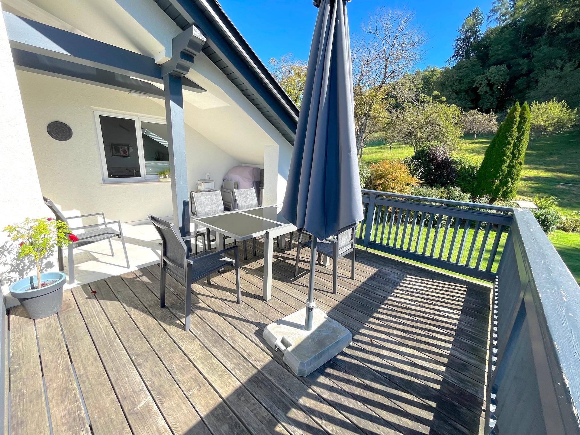 Sonnige Terrasse mit Holzboden, Esstisch und Sonnenschirm, umgeben von grüner Natur.