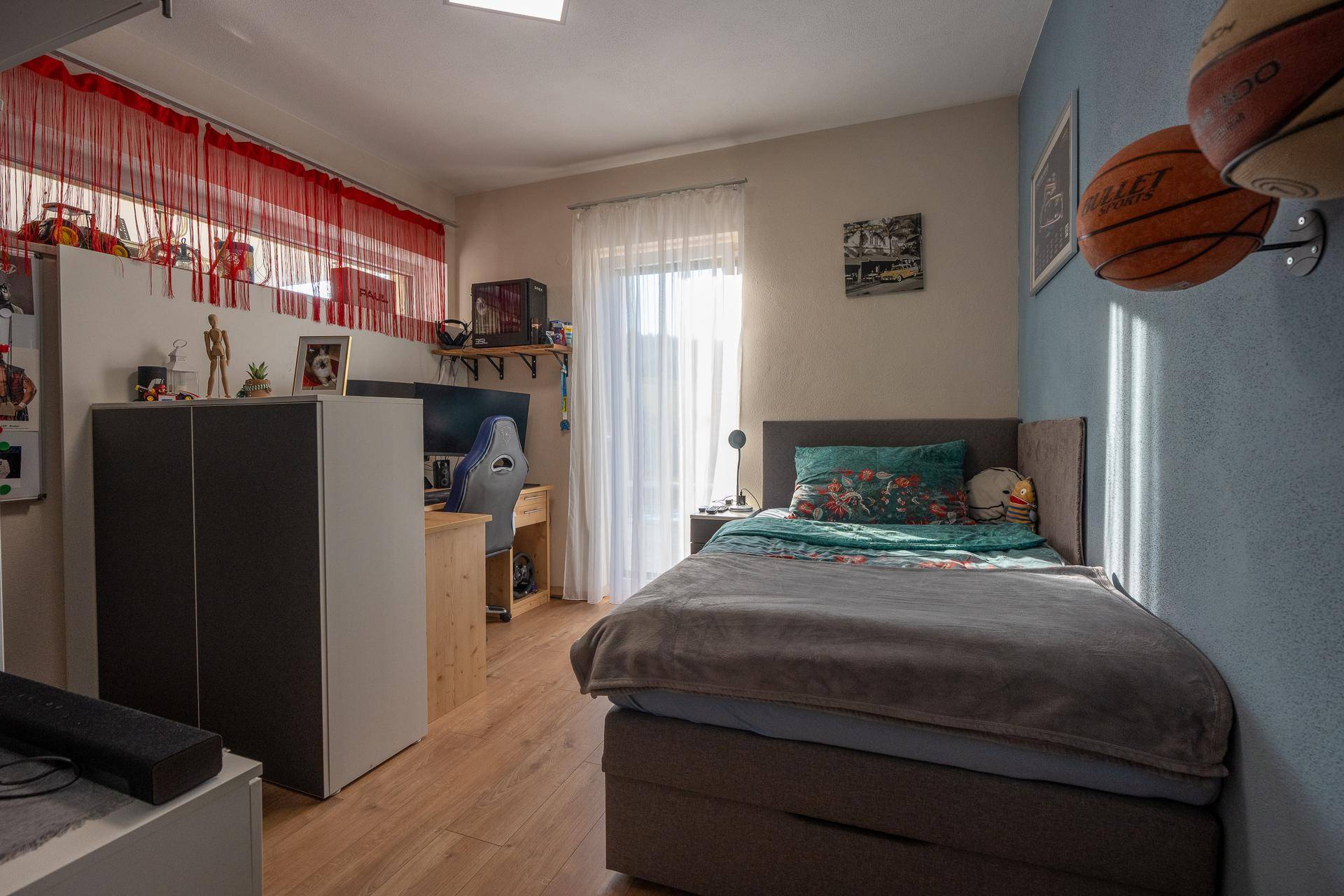 Jugendzimmer mit Einzelbett, Holzfußboden und Schreibtischbereich am Fenster.