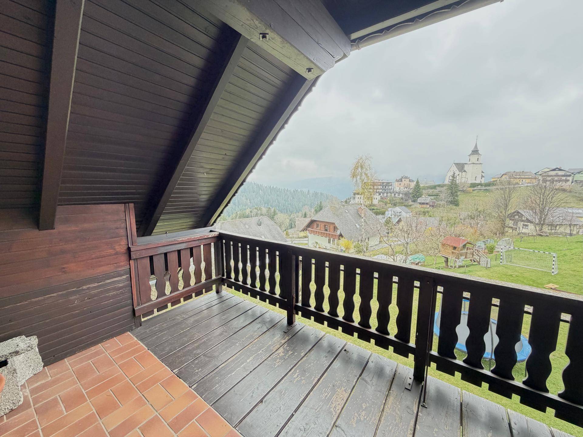 Rustikaler Holzbalkon mit weitem Blick über die Landschaft und ein Dorf mit Kirche.
