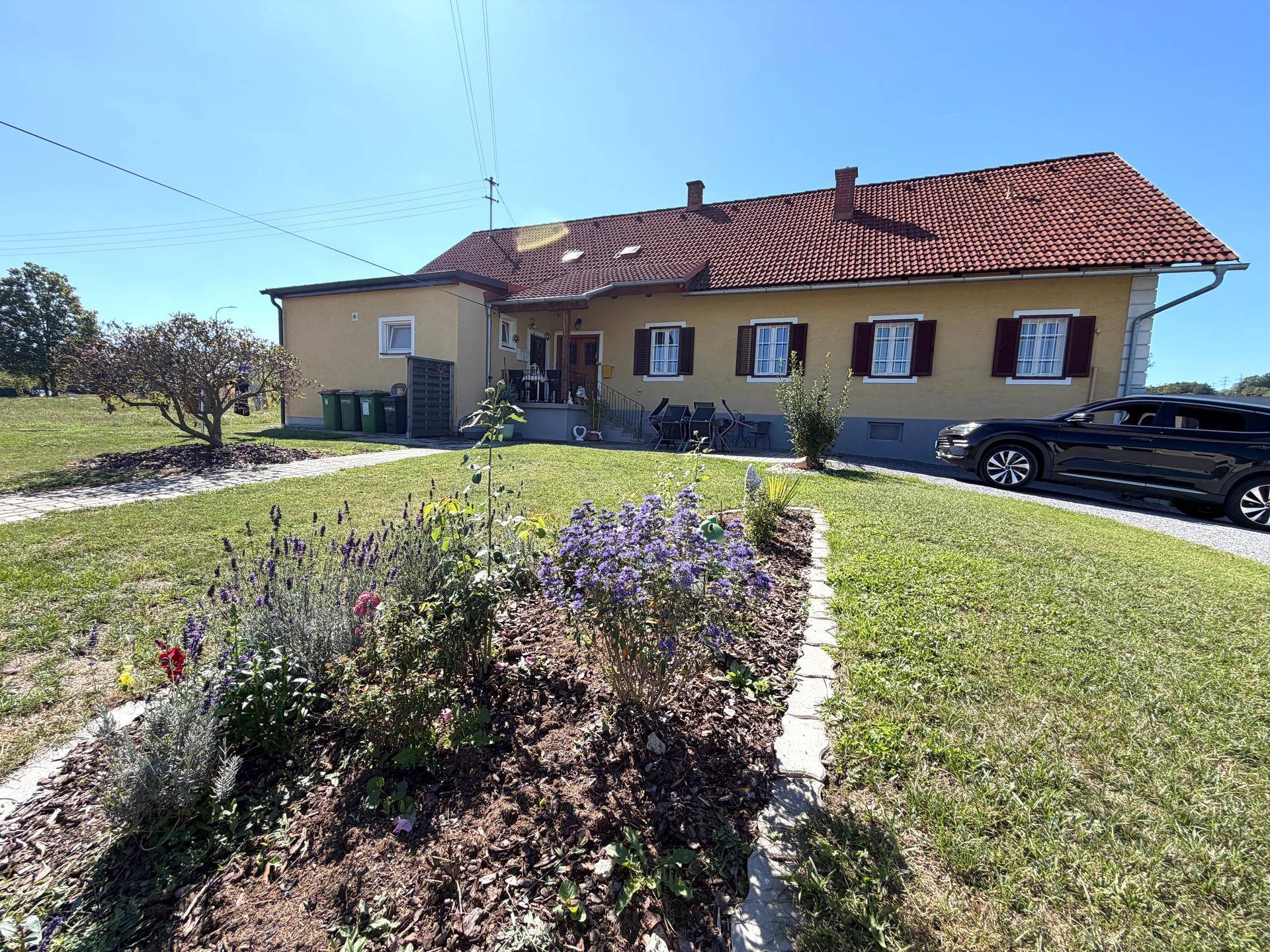 Vorderansicht des Hauses mit blühendem Blumenbeet und einer langen Fassade in ländlicher Umgebung.