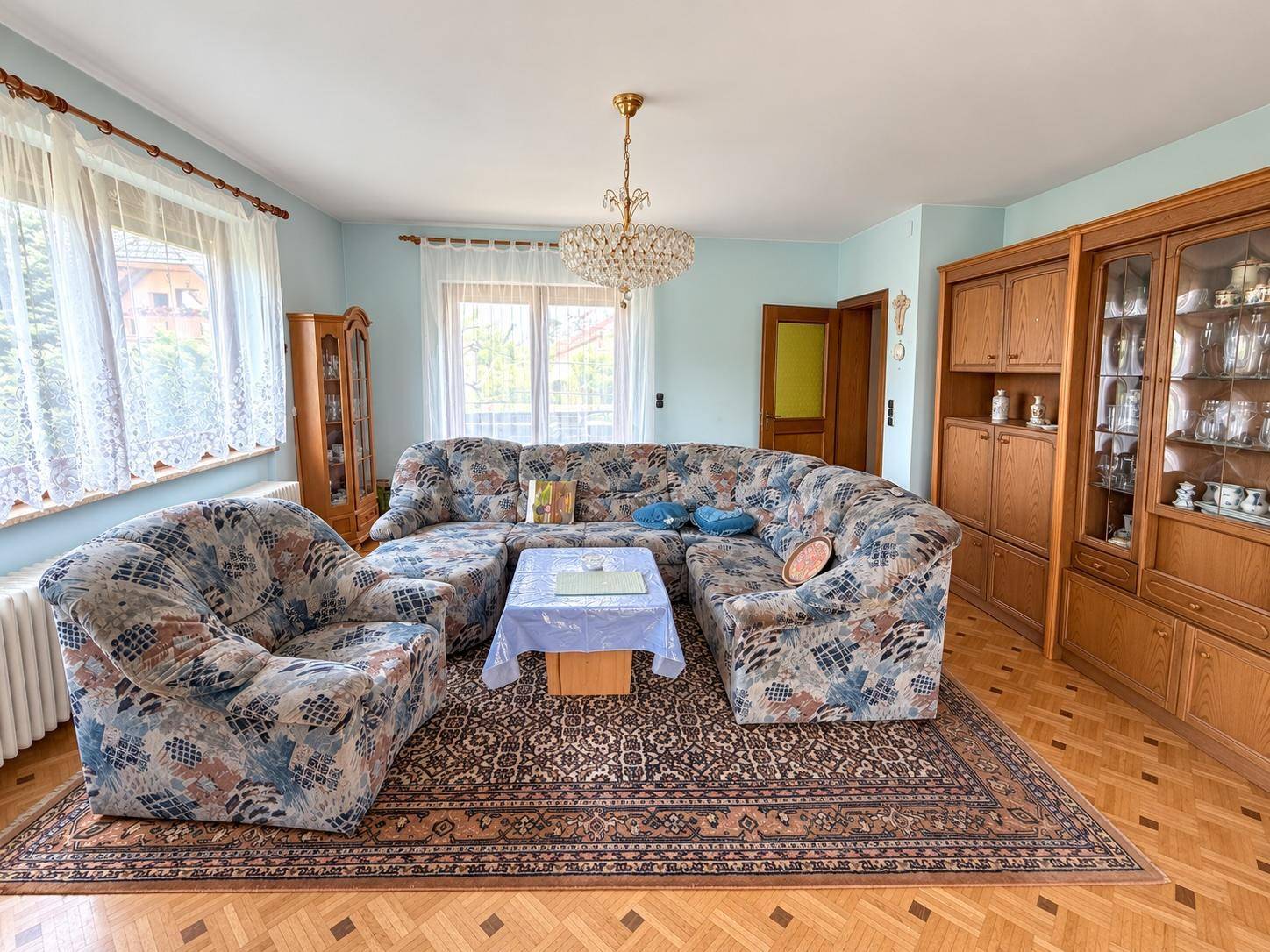 Helles Wohnzimmer mit gemustertem Sofa, Parkettboden und traditionellen Holzmöbeln.