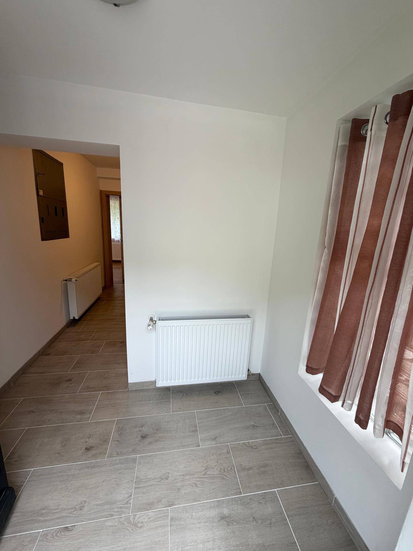 Heller Flur mit Fliesenboden, Heizkörper und Fenster mit Vorhang.