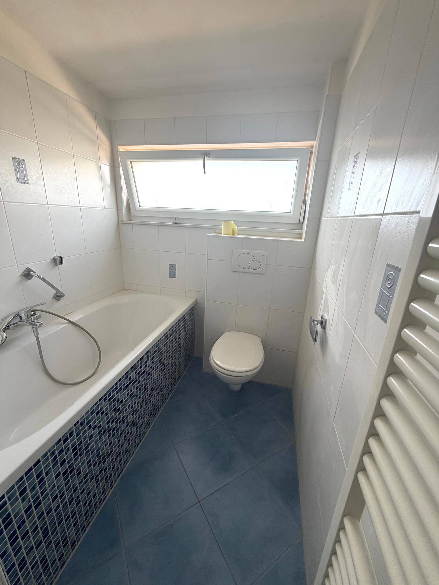Badezimmer mit Badewanne, WC, Fenster und blauen Bodenfliesen in gutem Zustand.