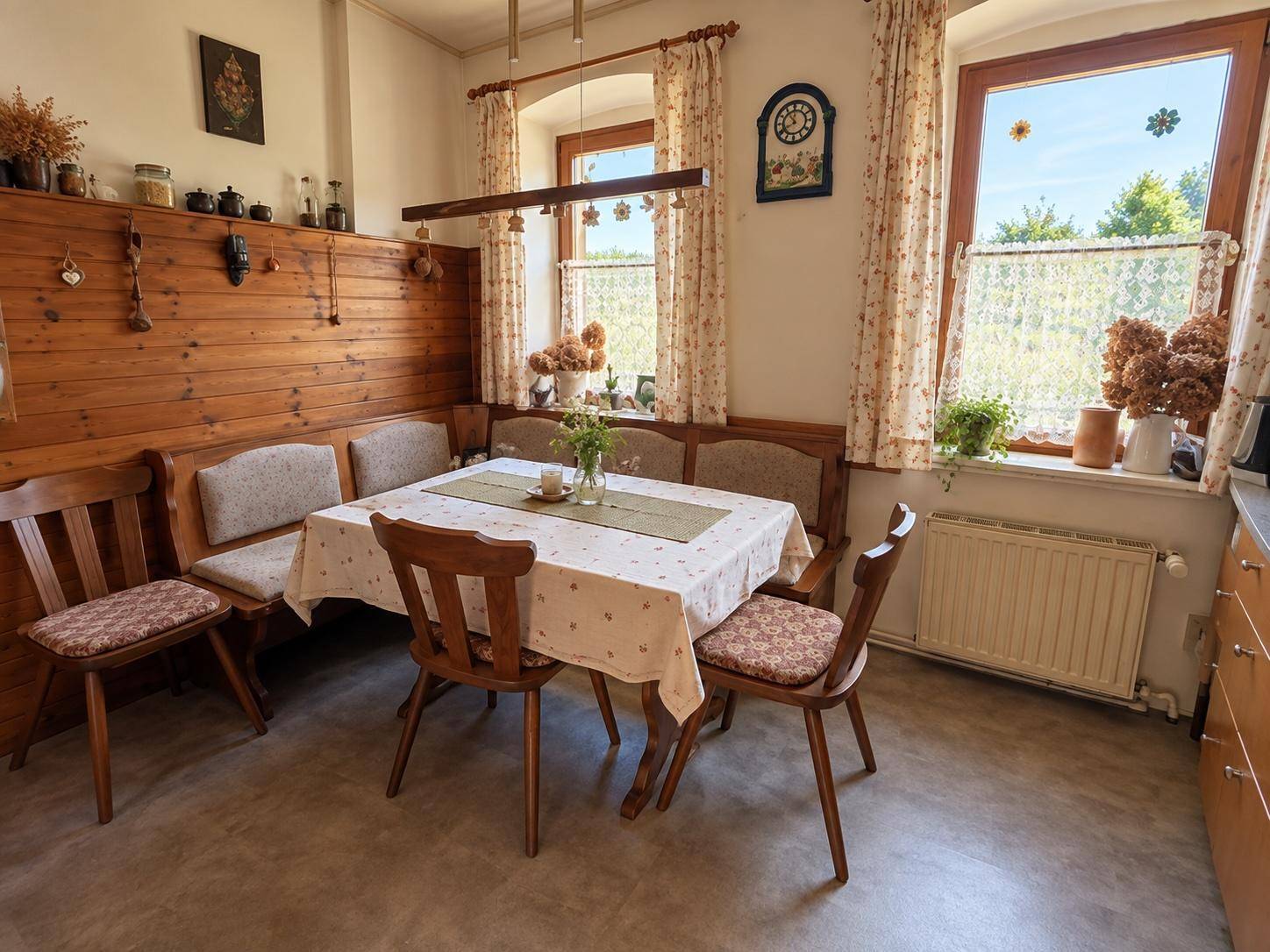 Detailansicht des Essbereichs in der Küche mit Holzbänken, Tisch und Vorhängen an den Fenstern.