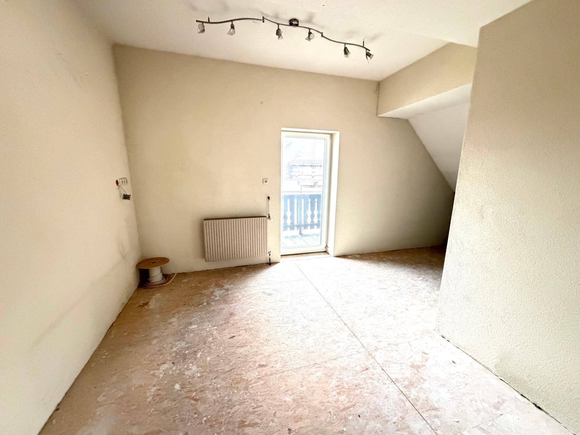 Dachgeschosszimmer im Ausbau mit schräger Decke, hellen Wänden und Zugang zu einem Außenbereich.