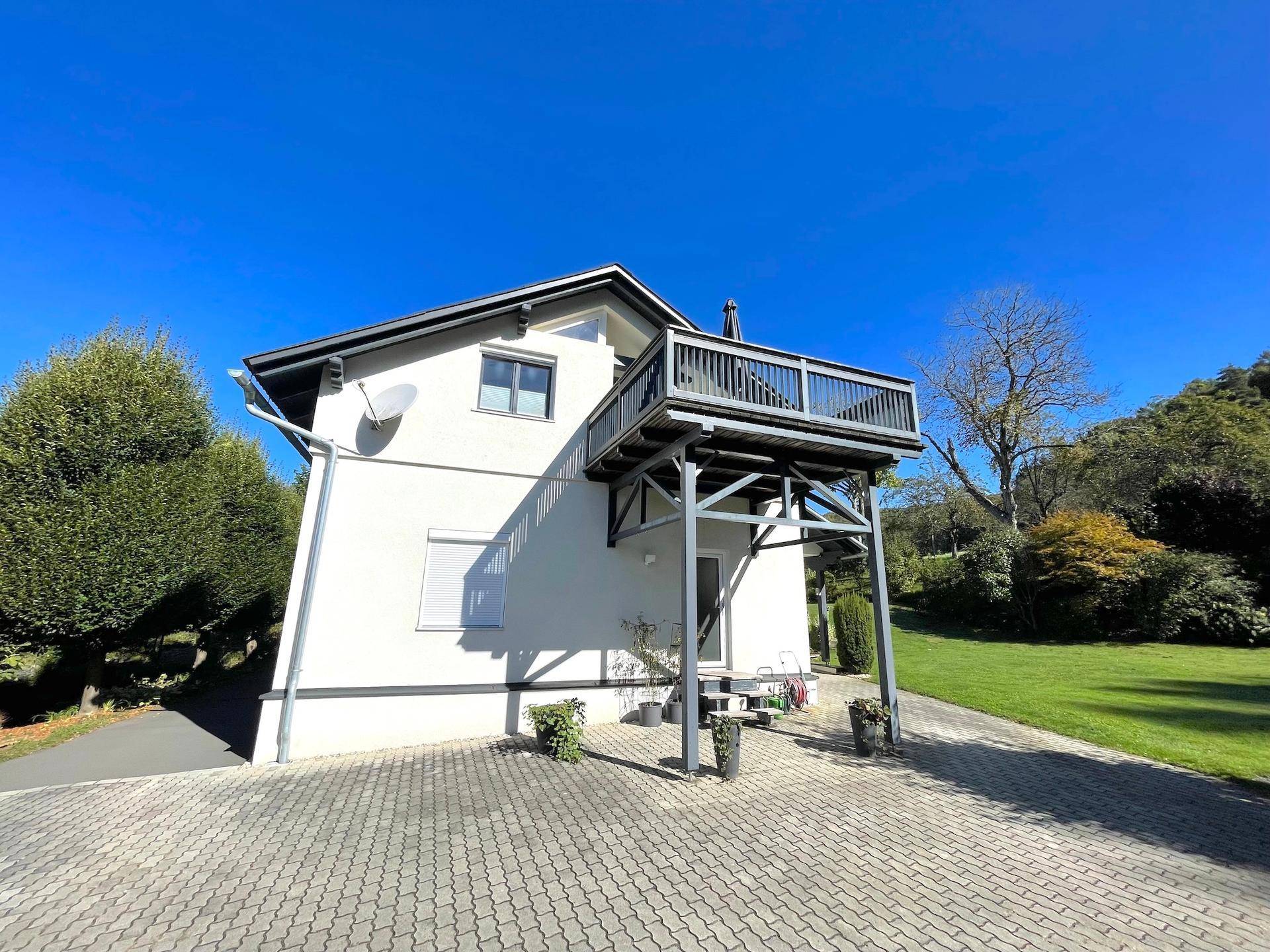 Seitenansicht des Hauses mit einem erhöhten Balkon auf Stützen und einem gepflasterten Vorplatz.