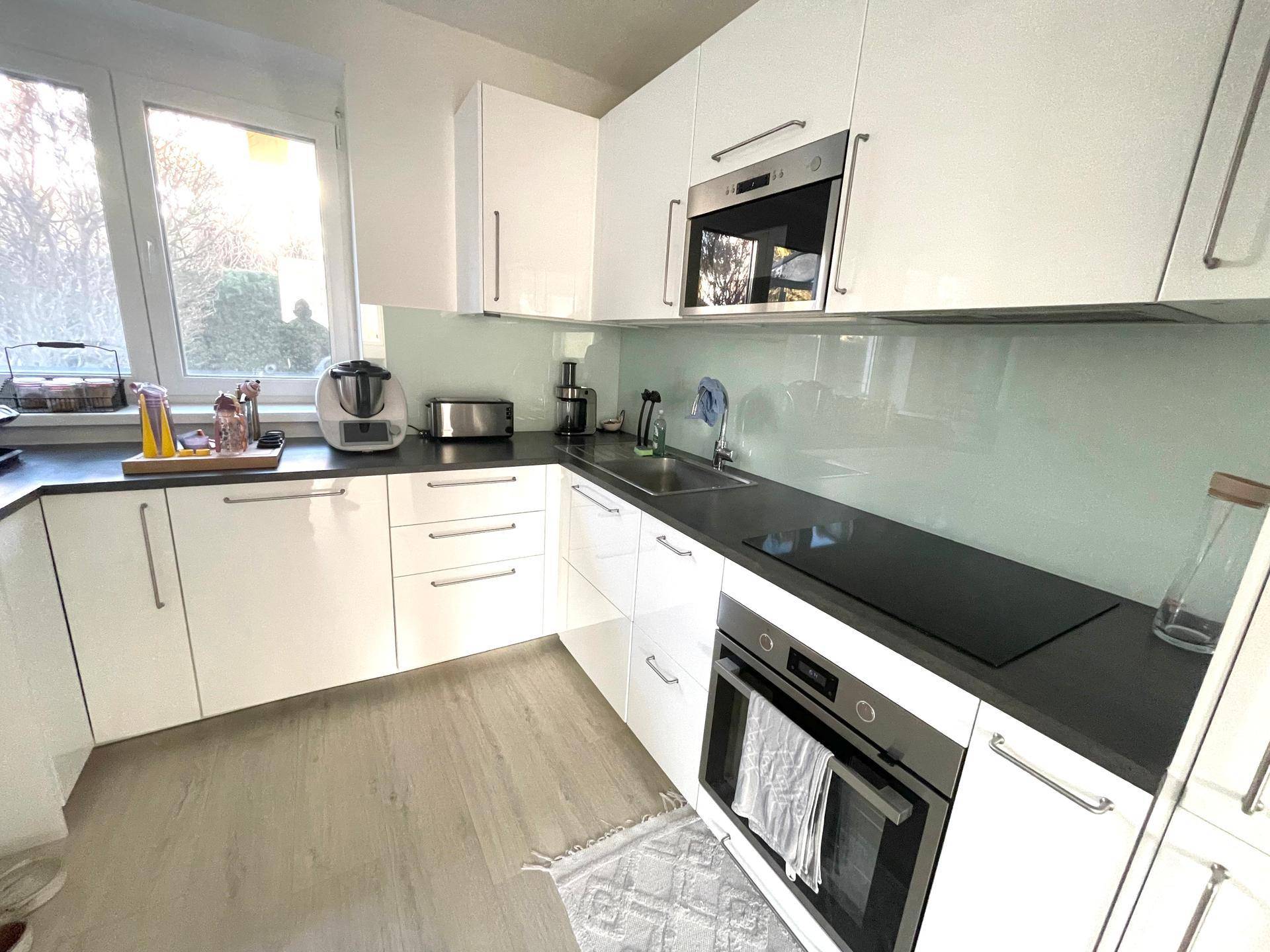 Moderne Küche mit weißen Hochglanzfronten, dunkler Arbeitsplatte und Fenster.