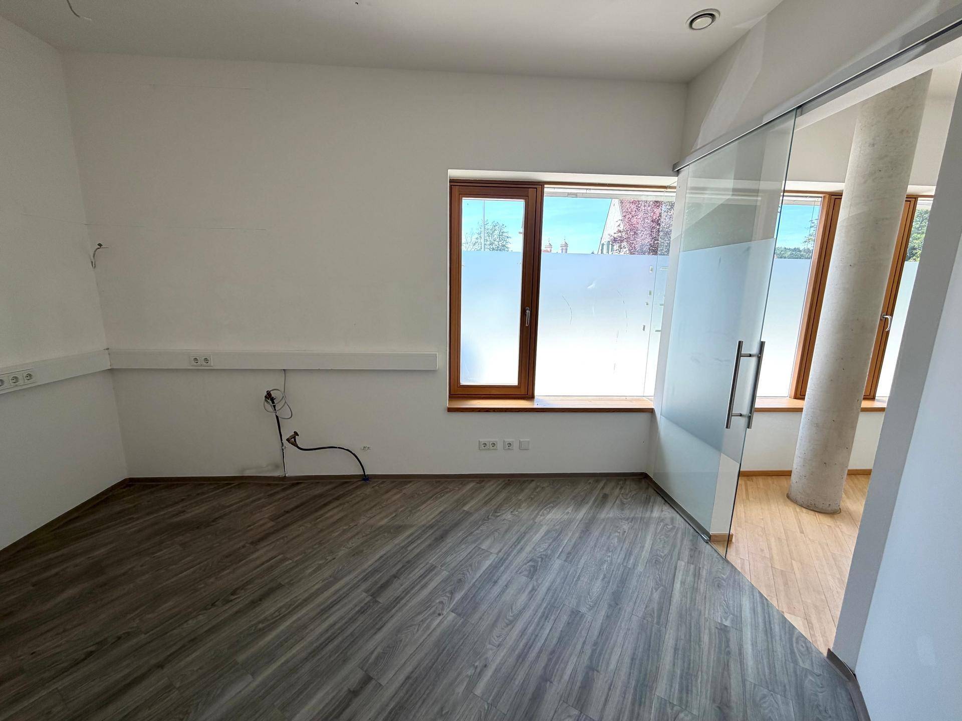 Geräumiges Büro mit grauem Laminatboden, großen Fenstern und einer modernen Glasschiebetür.