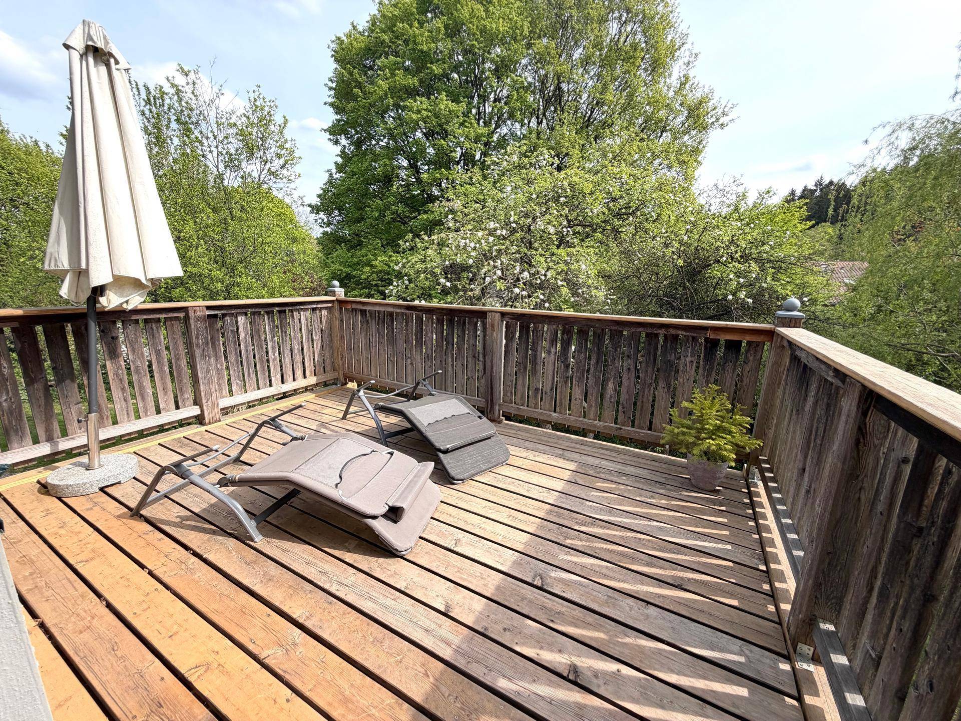 Großer Holzbalkon mit Sonnenliegen und Sonnenschirm, umgeben von üppigem Grün.