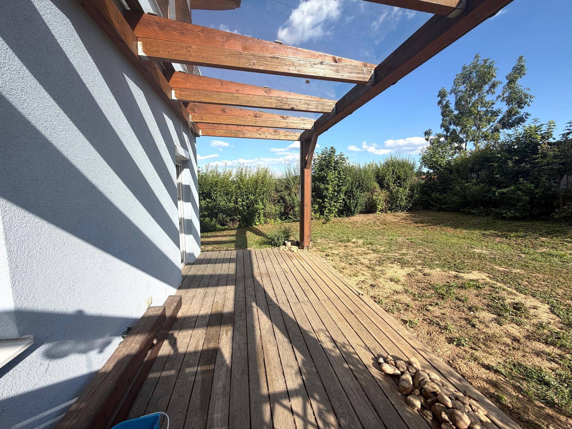 Holzterrasse mit Pergola und angrenzendem Gartenbereich unter strahlend blauem Himmel.