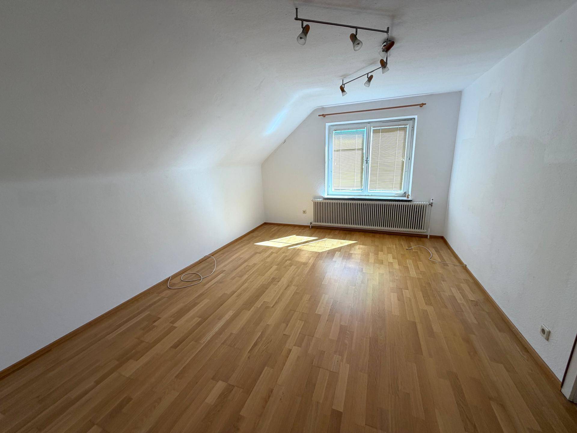 Helles Dachzimmer mit Holzboden, schrägen Wänden und einem Fenster mit Jalousien.