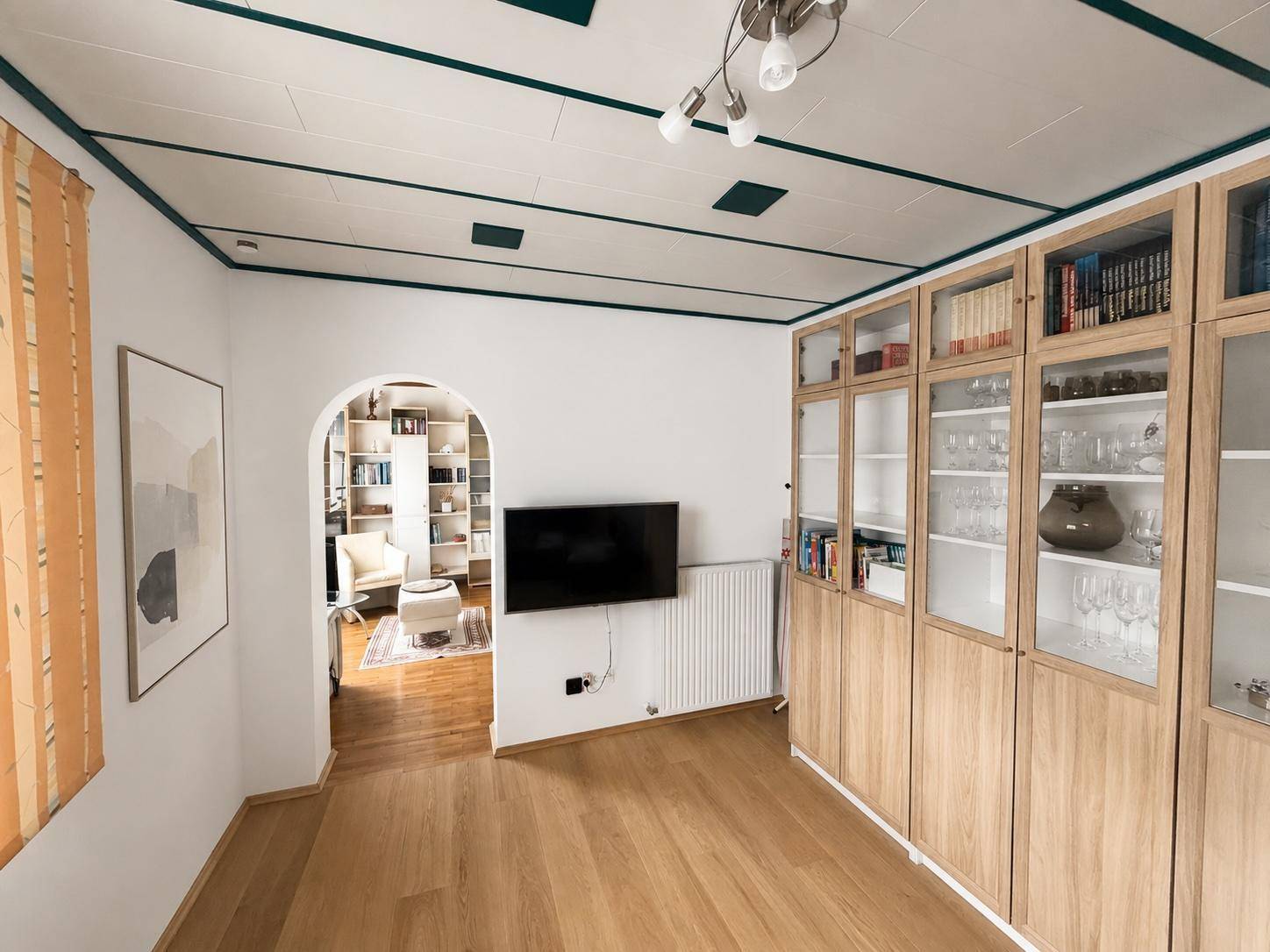 Wohnzimmer mit Laminatboden, Wandfernseher und Durchgang zu einem weiteren Raum.