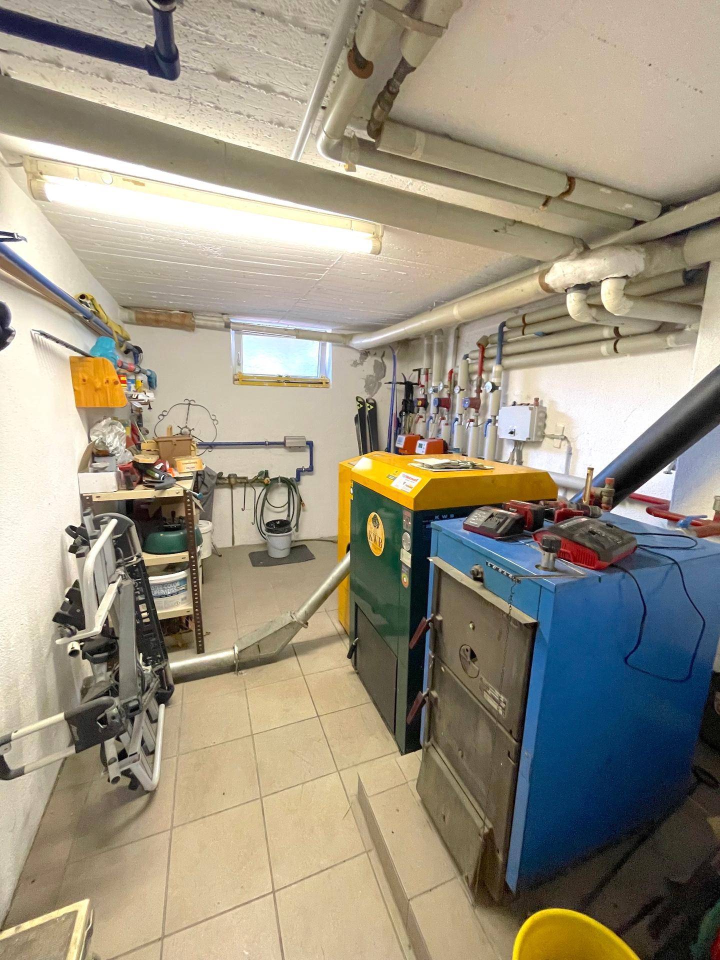 Heizungsraum im Keller mit zwei großen Heizkesseln, sichtbaren Rohren und einem kleinen Fenster.