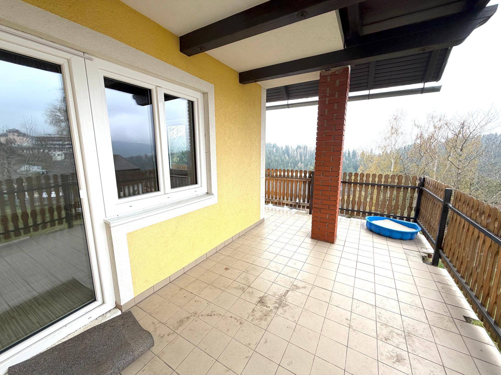 Überdachter Balkon mit Fliesenboden, traditionellem Holzgeländer und Blick auf die bewaldete Umgebung.