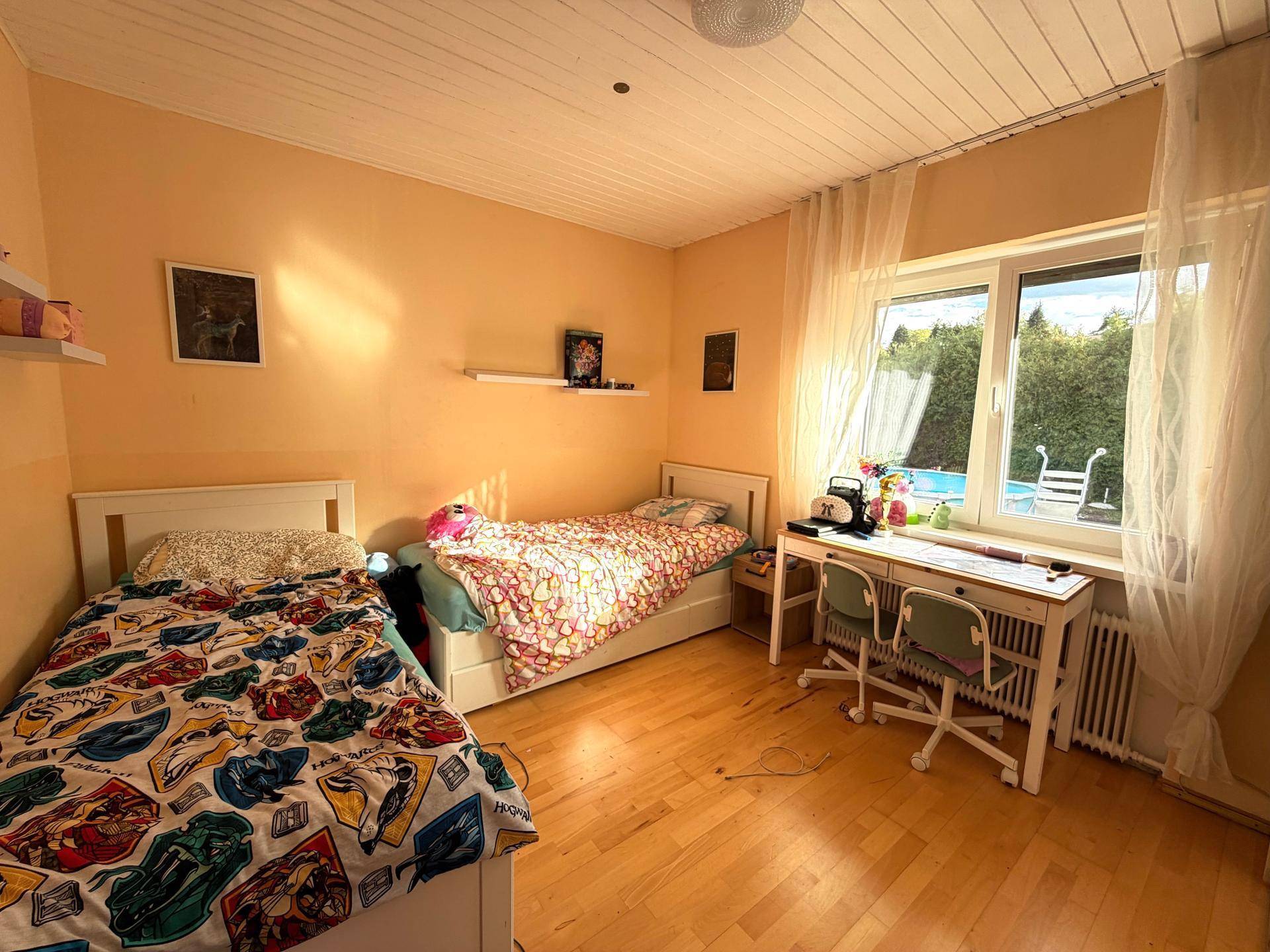 Helles Kinderzimmer mit zwei Betten, Schreibtisch und Blick auf den Garten und Pool.