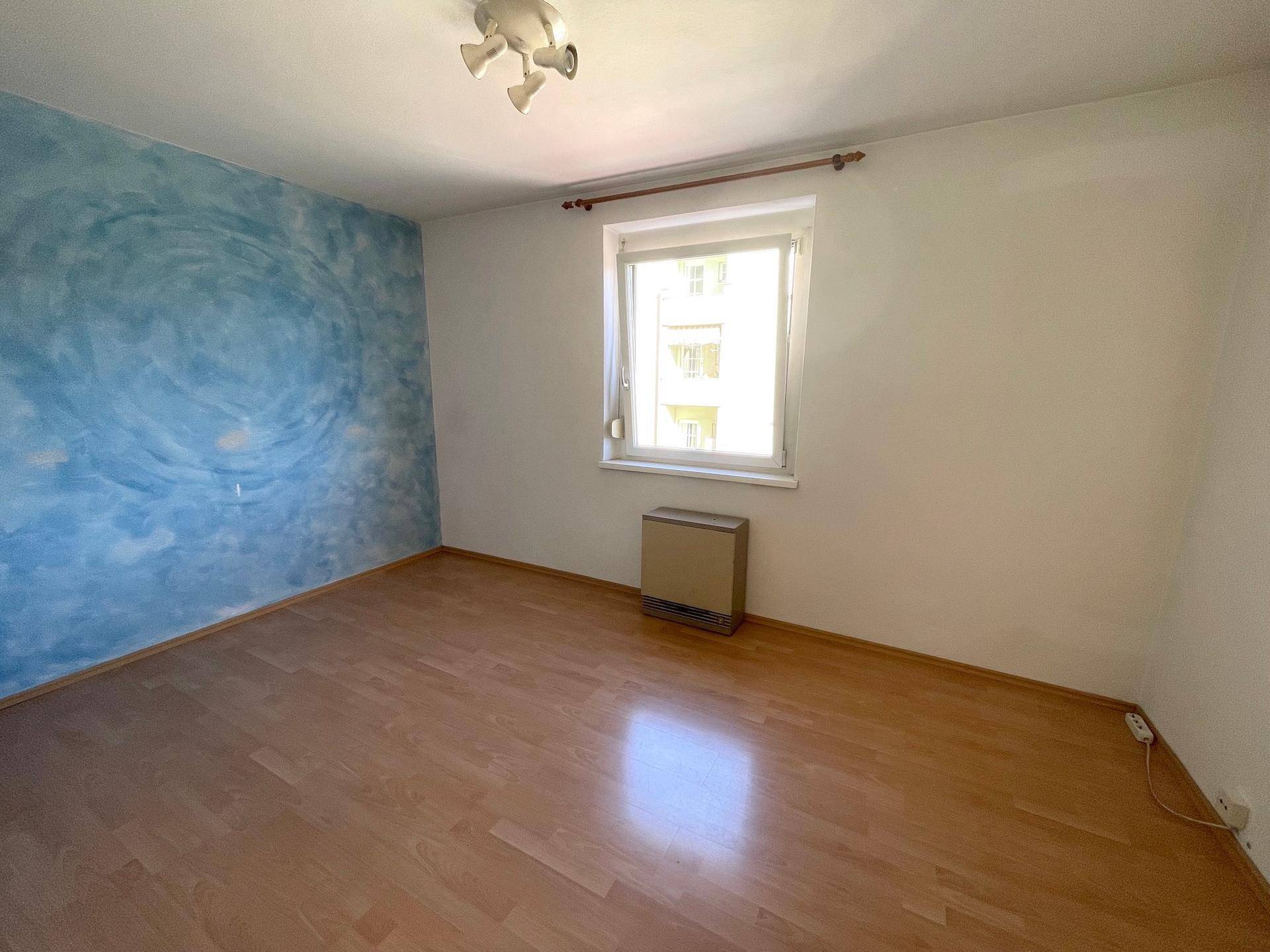 Schlafzimmer mit Parkettboden, blauer Akzentwand und großem Fenster.