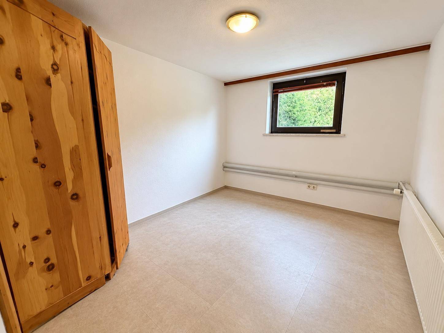 Kleines Schlafzimmer mit hellem Fliesenboden, weiß gestrichenen Wänden und Holzschrank.