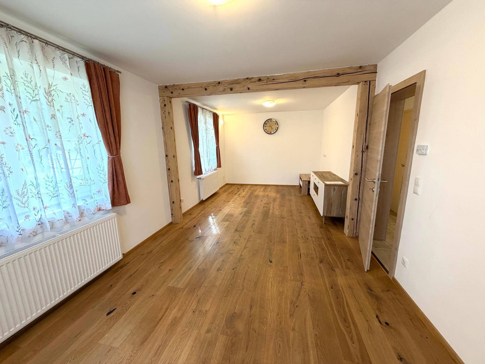 Geräumiges Wohnzimmer mit Holzbalken, Parkettboden und großen Fenstern mit Vorhängen.
