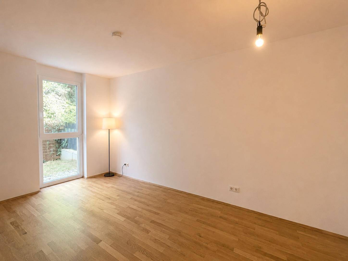 Lichtdurchfluteter Raum mit Holzfußboden und bodentiefem Fenster, das den Blick ins Grüne freigibt.