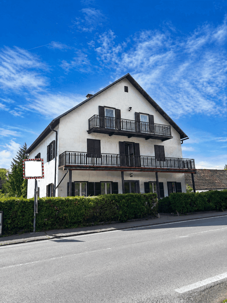 Frontansicht des Wohnhauses mit Balkonen, Fensterläden und einer grünen Hecke unter blauem Himmel.