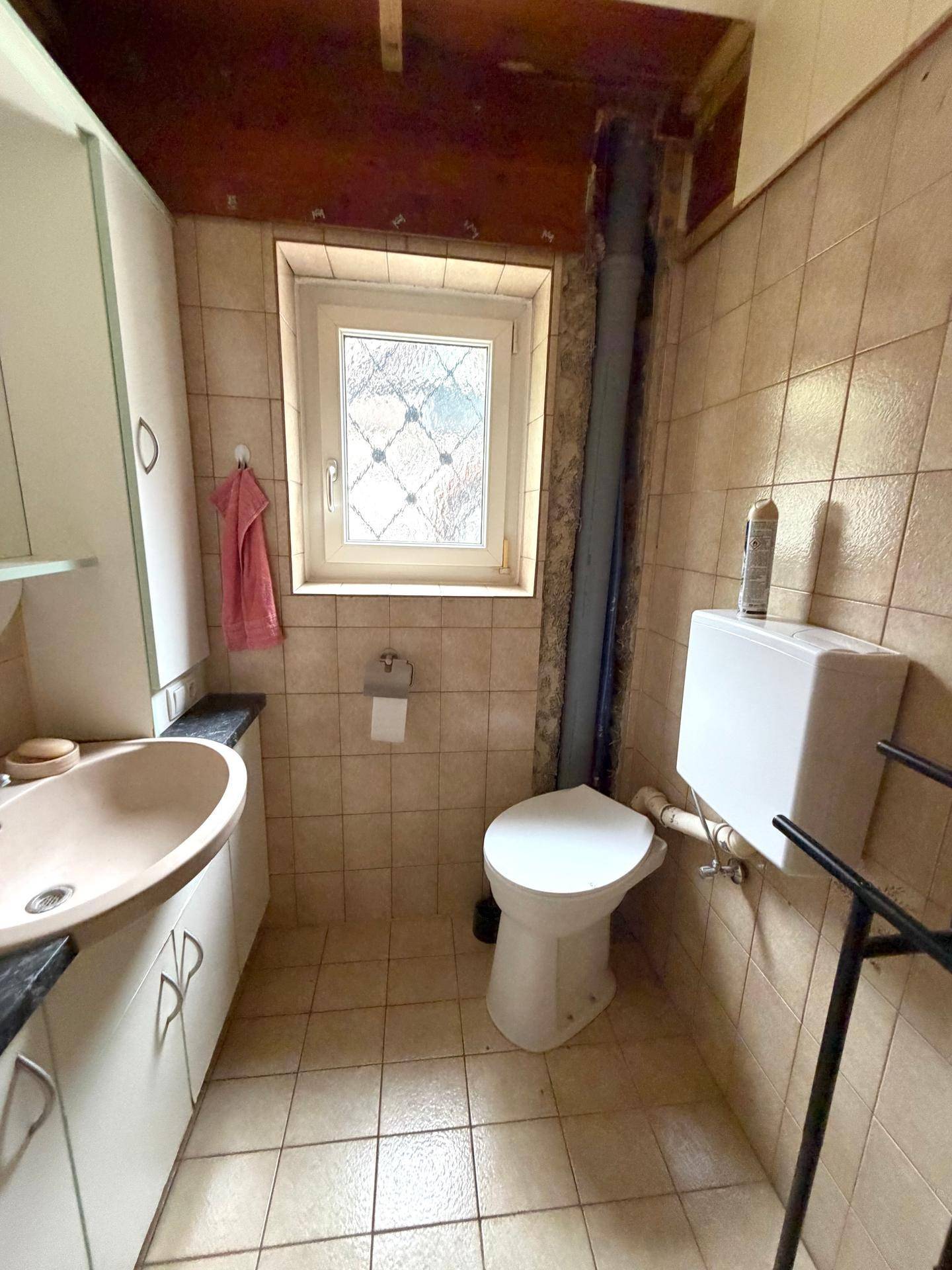 Kleines WC mit Fenster, Waschtisch und Toilette, teilweise mit unverputzter Decke.