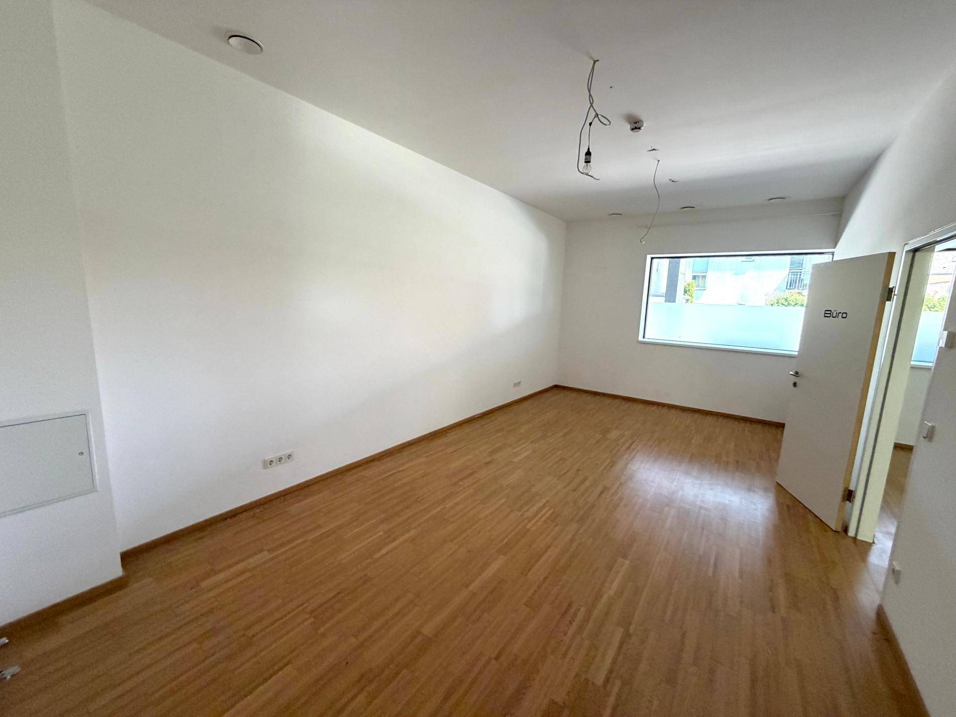 Leeres Büro mit hellem Holzfußboden, weißen Wänden und einem Fenster mit Sichtschutzfolie.
