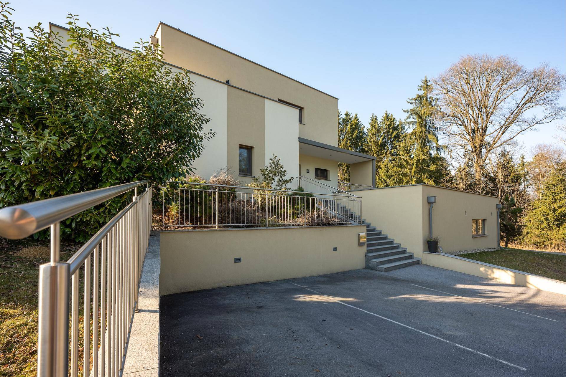 Ansicht der modernen Villa mit Treppenaufgang, Metallgeländer und umgebender Bepflanzung.